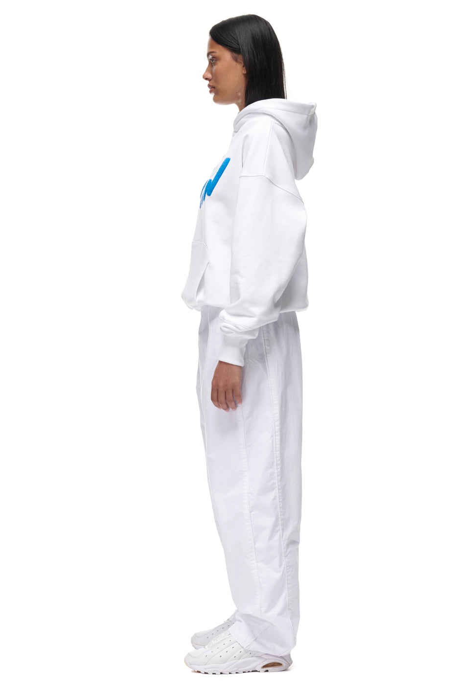 A woman stands in profile against a plain white background, wearing the 6PM PLAY HOODIE WHITE with matching white pants and sneakers.