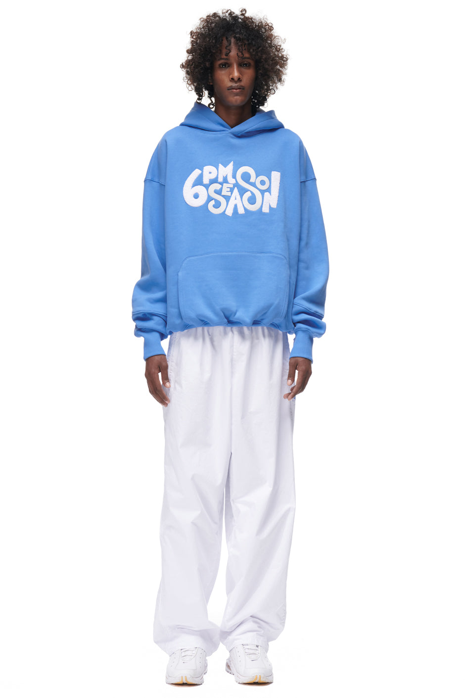 A person faces forward wearing the PLAY HOODIE BLUE by 6PM, featuring bold white "6PM SEASON" text, paired with baggy white pants and white sneakers, all set against a plain white background.