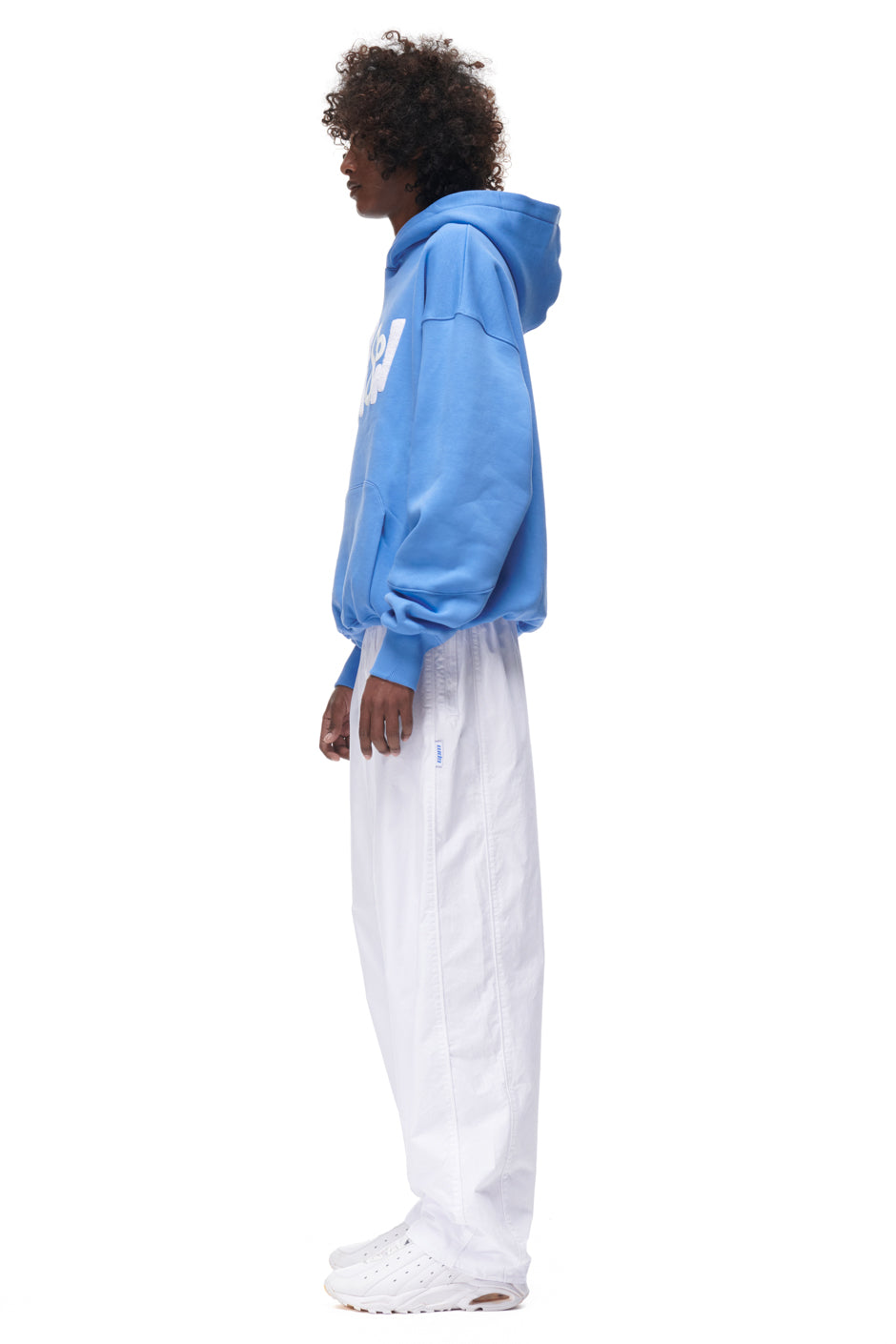 A person with curly hair stands in profile, wearing the 6PM PLAY HOODIE BLUE, loose white pants, and white sneakers, on a plain white background.