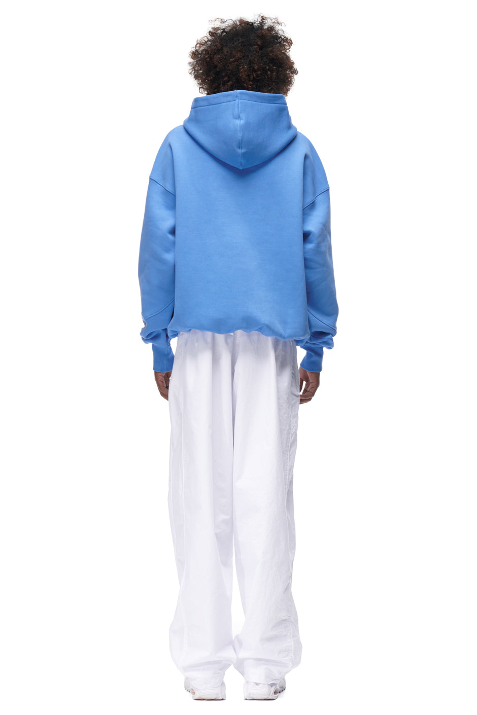 A person faces away from the camera wearing the 6PM PLAY HOODIE BLUE with the hood up and oversized white pants; hands and feet visible against a plain white background.