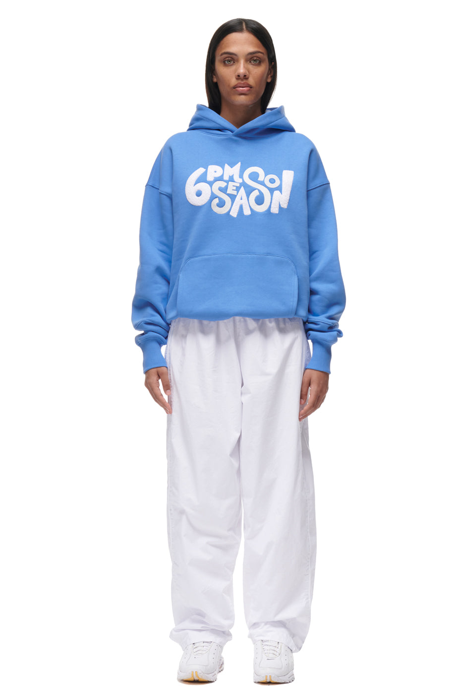 A woman with straight dark hair and a neutral expression wears the 6PM PLAY HOODIE BLUE, featuring bold white "6PM SEASON" lettering, paired with loose white pants, standing against a plain white background.