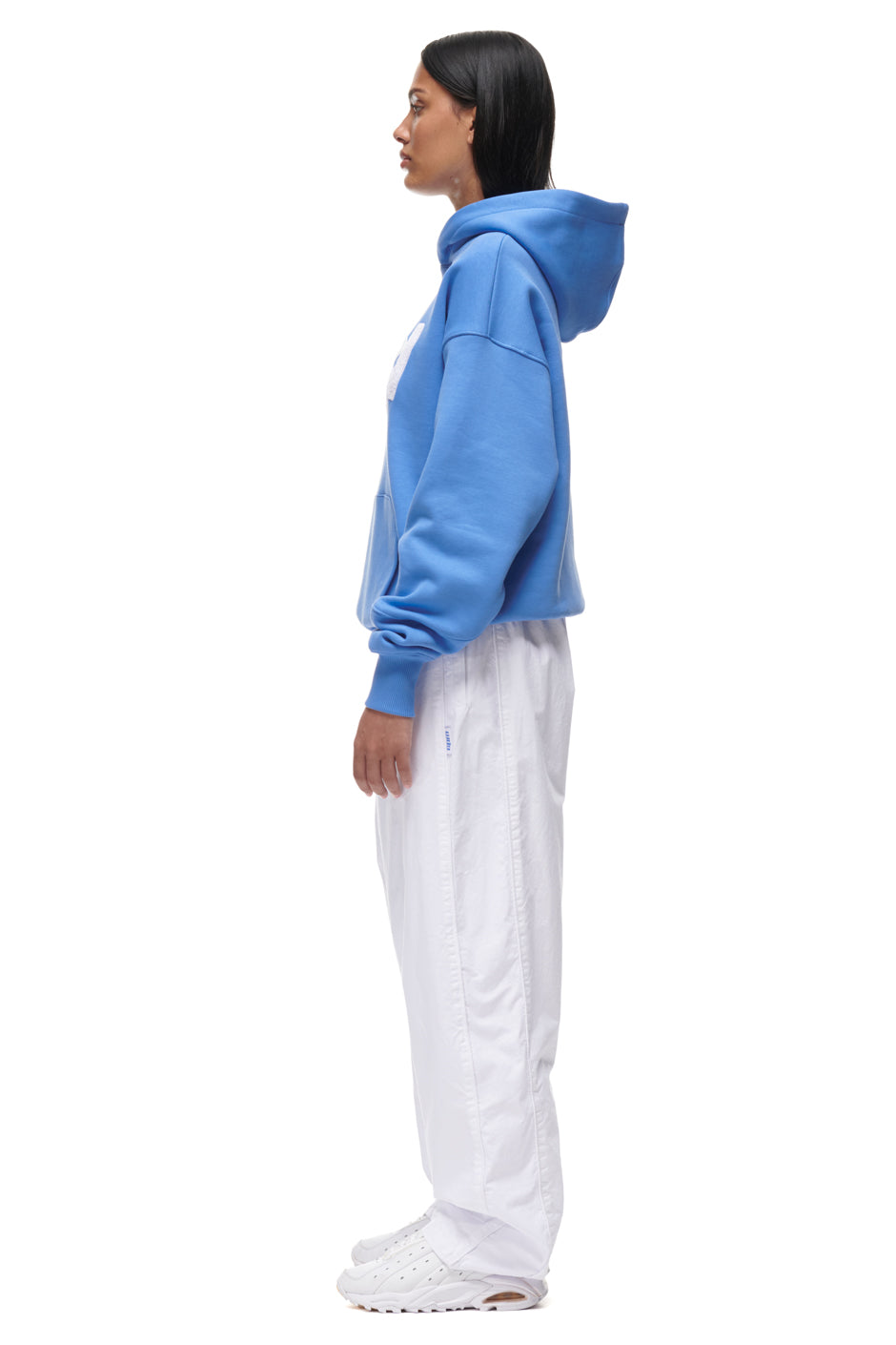 A person stands in profile against a plain white background, wearing the 6PM PLAY HOODIE BLUE, paired with white pants and white sneakers.