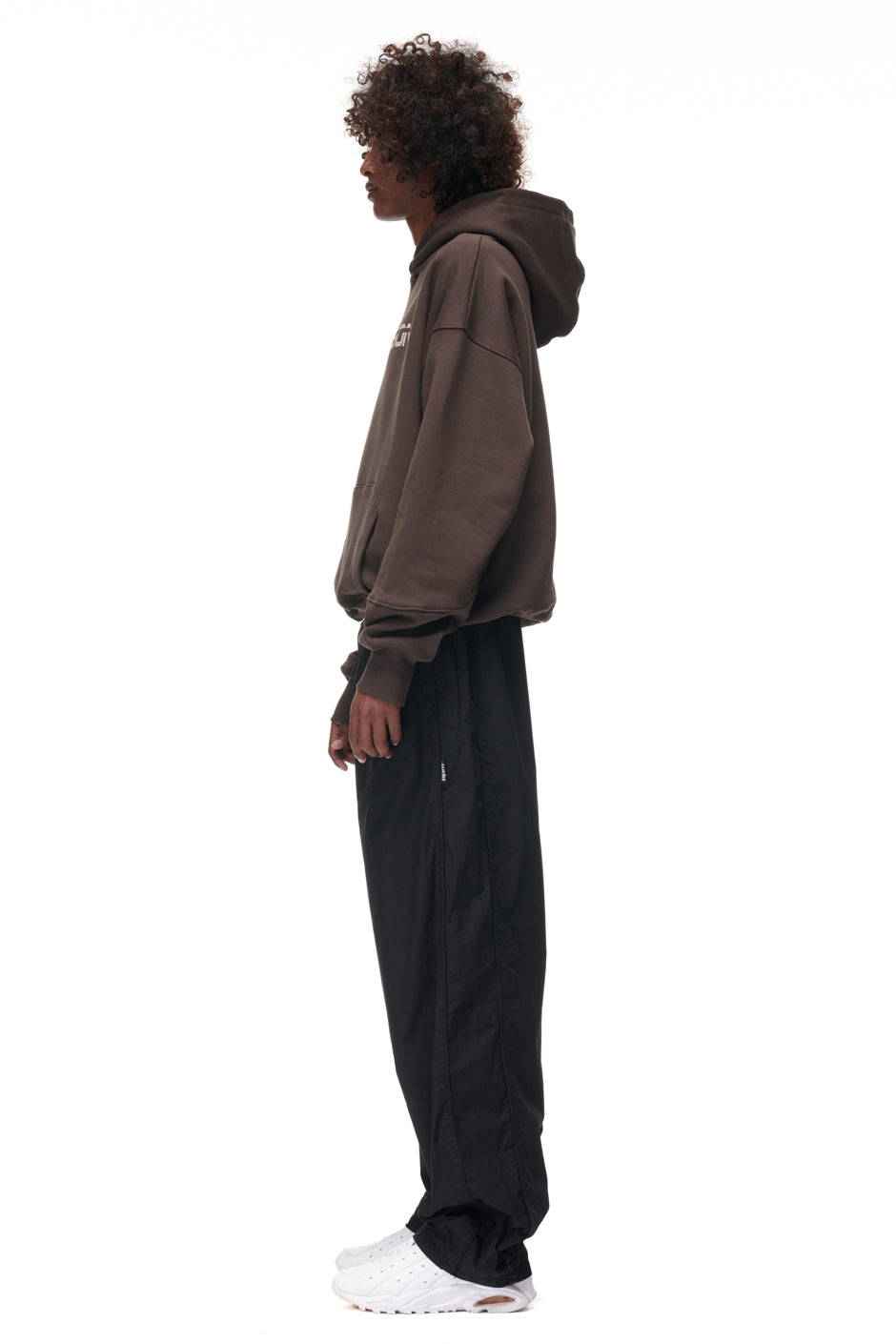 A person stands in profile against a white background, wearing the 6PM TILES HOODIE BROWN, loose black pants, and white sneakers.