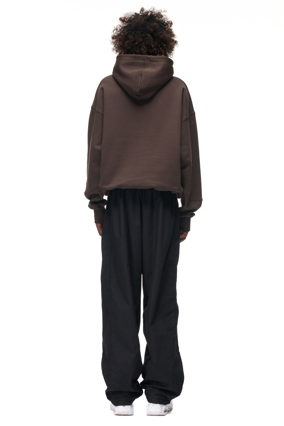 A person with curly hair faces away, wearing the 6PM TILES HOODIE BROWN, loose black pants, and white shoes against a plain white background.