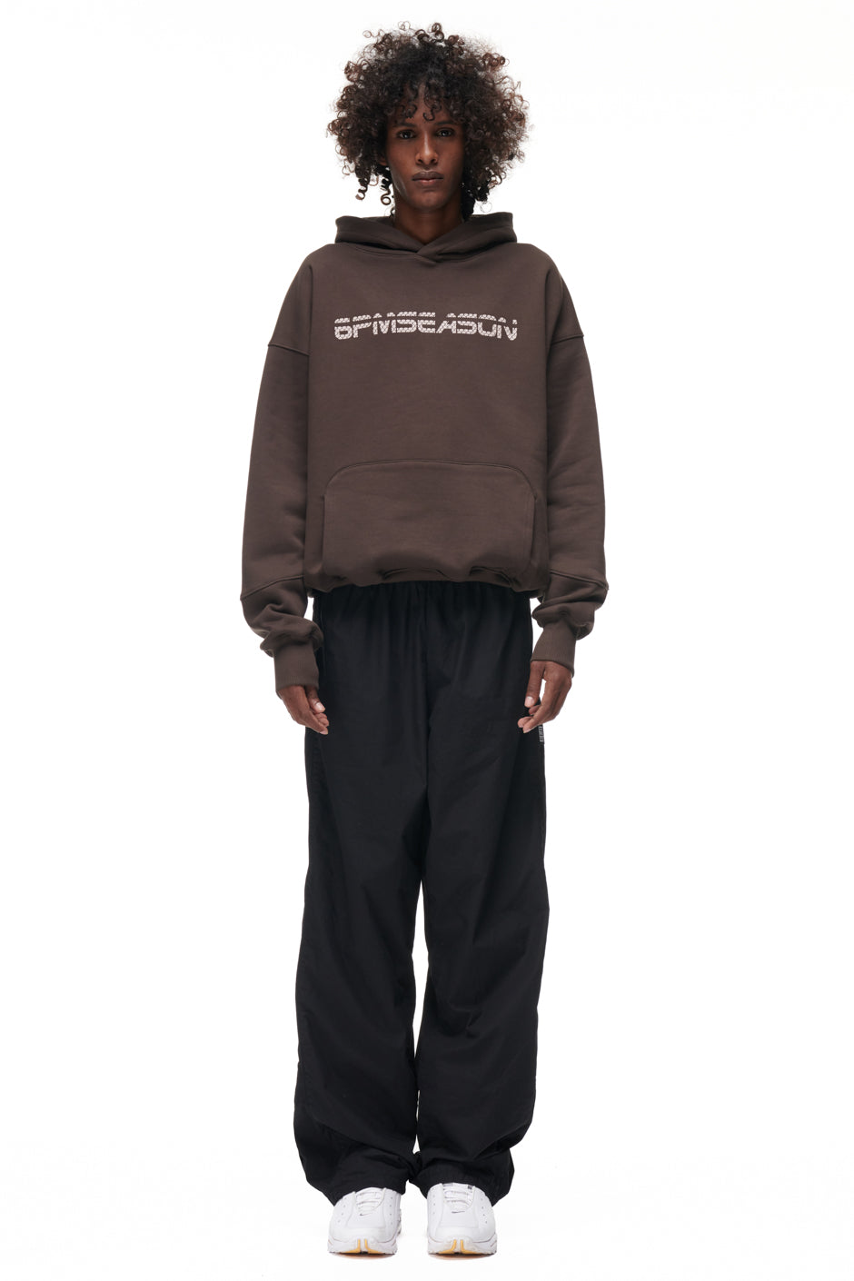 A person with curly hair faces forward, wearing the 6PM TILES HOODIE BROWN, loose black pants, and white sneakers against a plain white background.