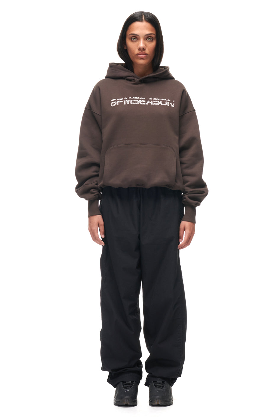 A woman with straight, shoulder-length hair and a neutral expression faces forward, wearing the 6PM TILES HOODIE BROWN, black loose pants, and black sneakers against a white background.