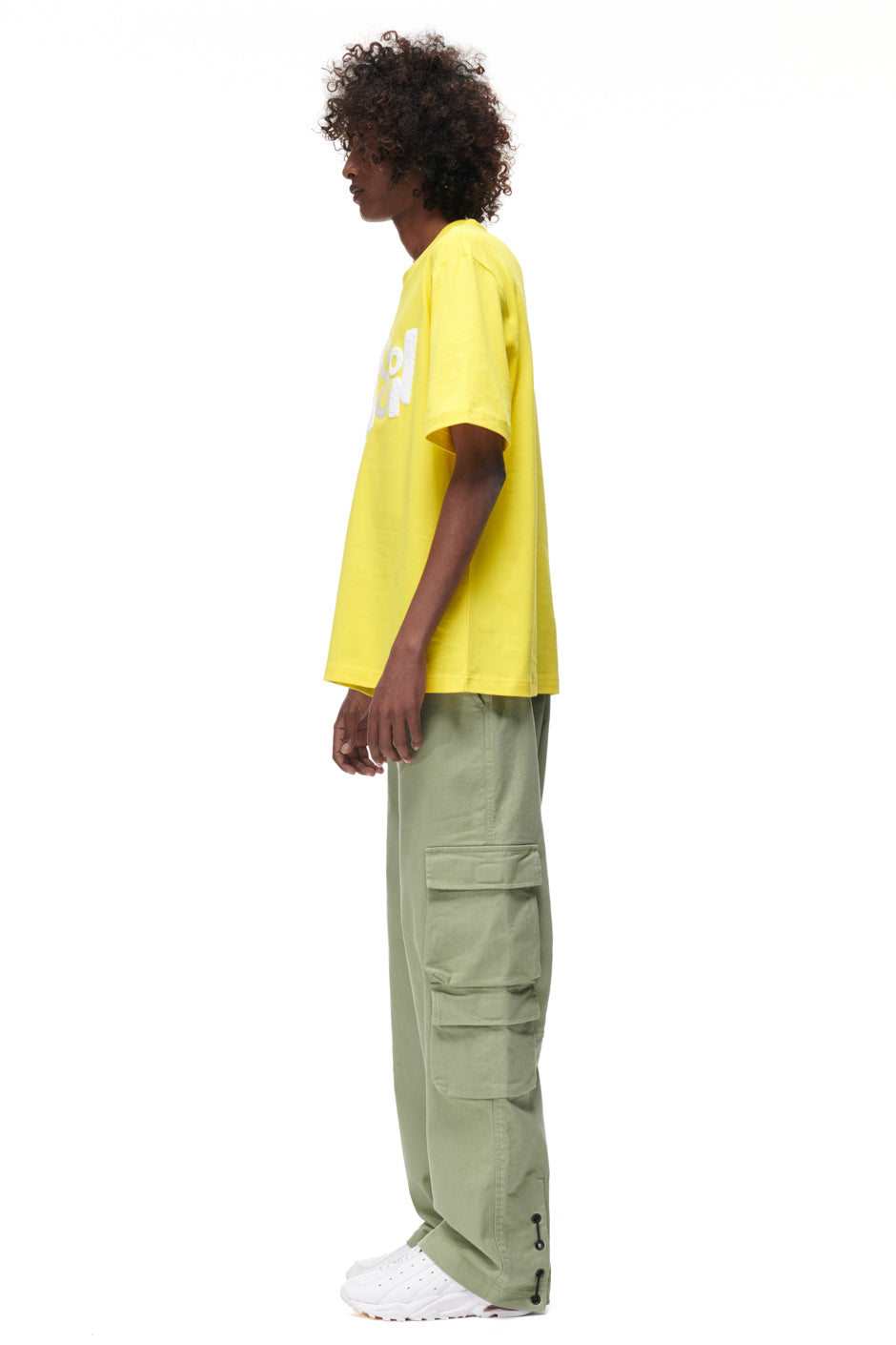 A person with curly hair stands in profile, wearing the PLAY T-SHIRT YELLOW by 6PM, light green cargo pants with large side pockets, and white sneakers against a plain white background.