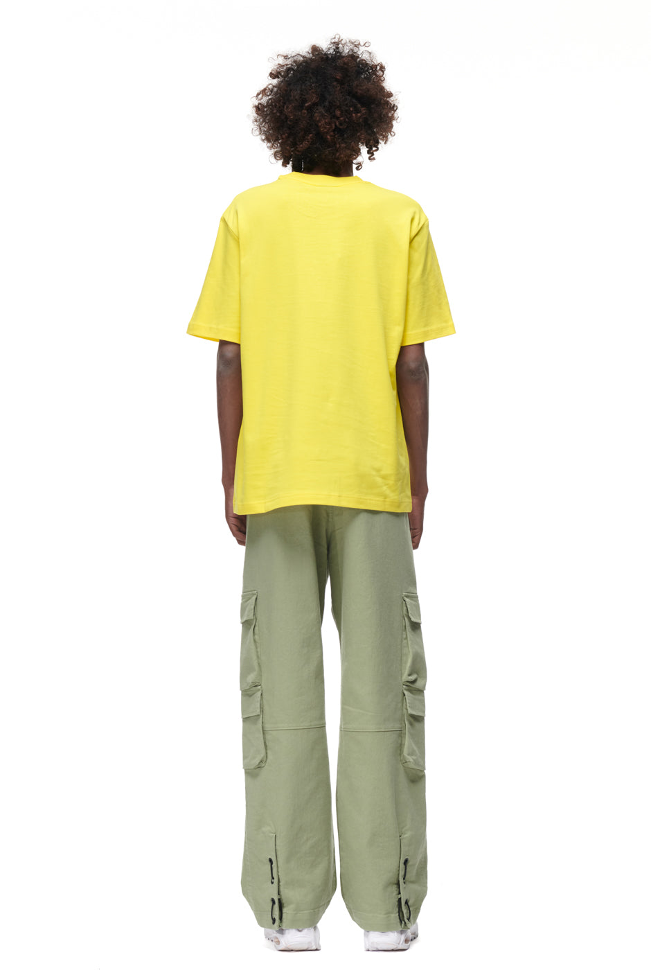 A person with curly hair faces away, wearing the 6PM PLAY T-SHIRT YELLOW and light green cargo pants with side pockets against a plain white background.