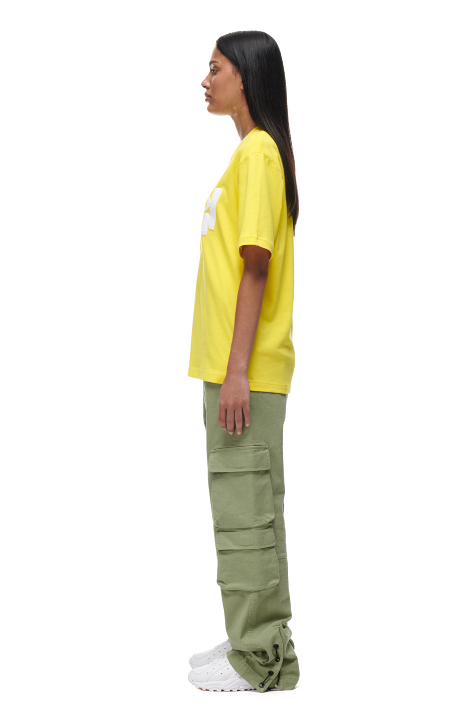 A person with long dark hair stands in profile against a white background, wearing the 6PM PLAY T-SHIRT YELLOW, olive green cargo pants with large pockets, and white sneakers.