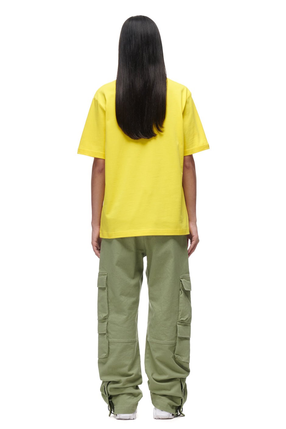 A person with long dark hair faces away in a 6PM PLAY T-SHIRT YELLOW, olive green cargo pants with side pockets, and white shoes, all set against a plain white background.