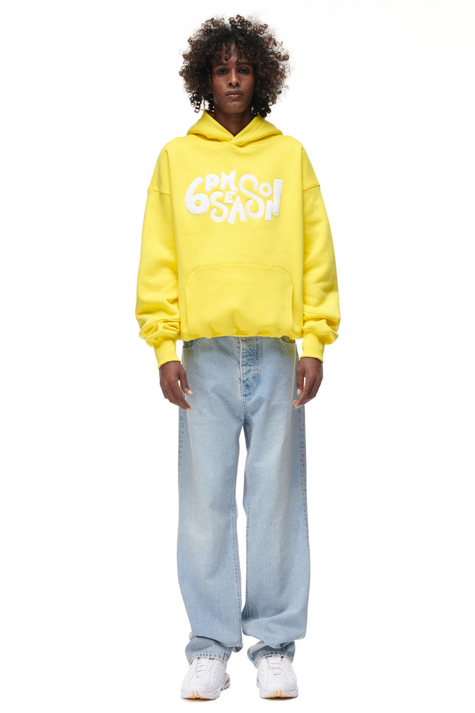 A person stands facing forward wearing the 6PM PLAY HOODIE YELLOW, light blue baggy jeans, and white sneakers against a plain white background.