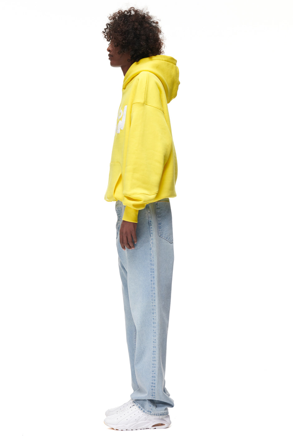 A person stands in profile against a plain white background wearing the 6PM PLAY HOODIE YELLOW, paired with light blue jeans and white sneakers.