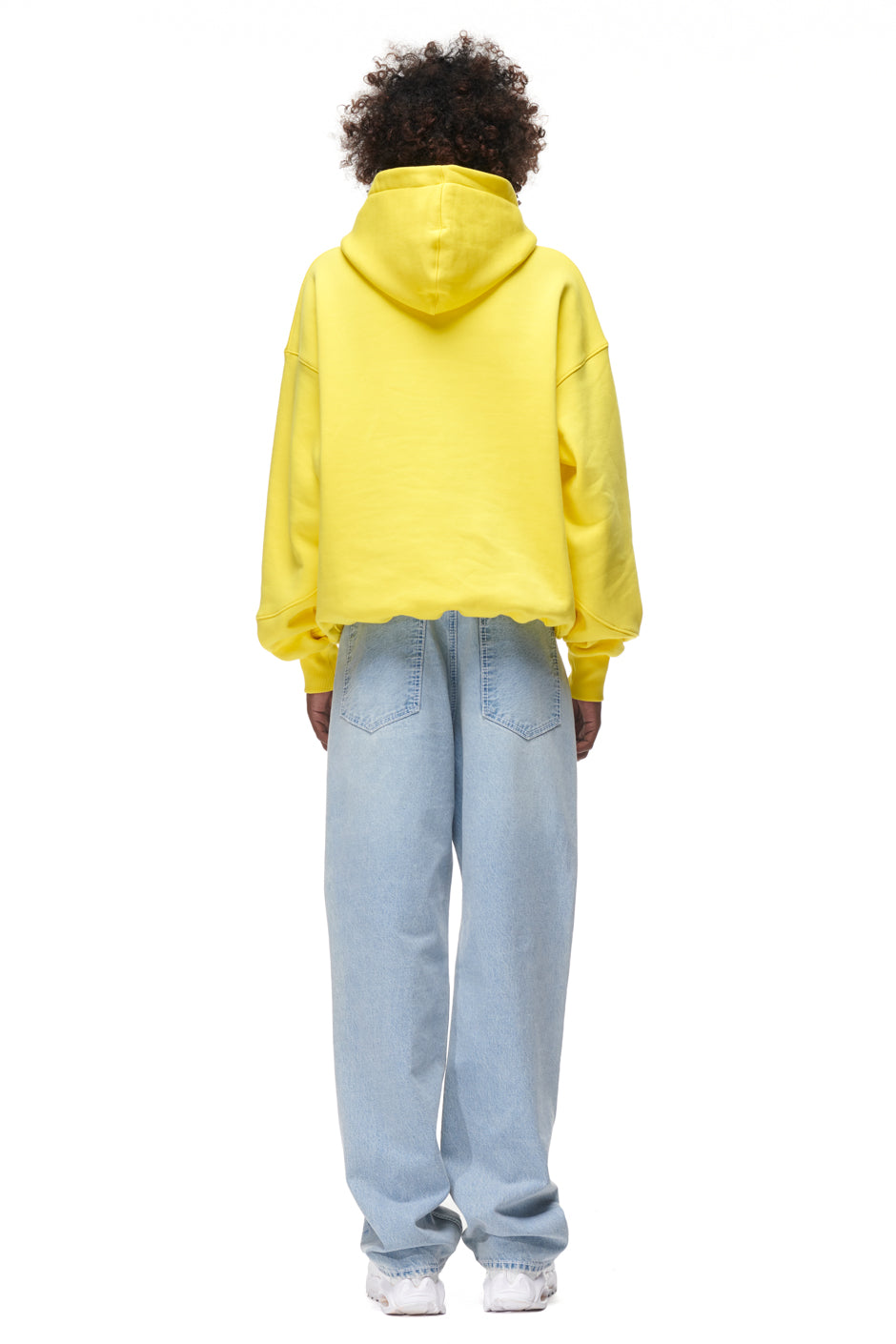 A person stands with their back to the camera, wearing the 6PM PLAY HOODIE YELLOW, light blue loose-fit jeans, and white sneakers against a plain white background.
