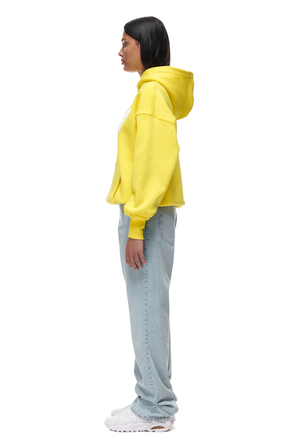 A woman stands in profile against a white background, wearing the 6PM PLAY HOODIE YELLOW, light blue jeans, and white sneakers.
