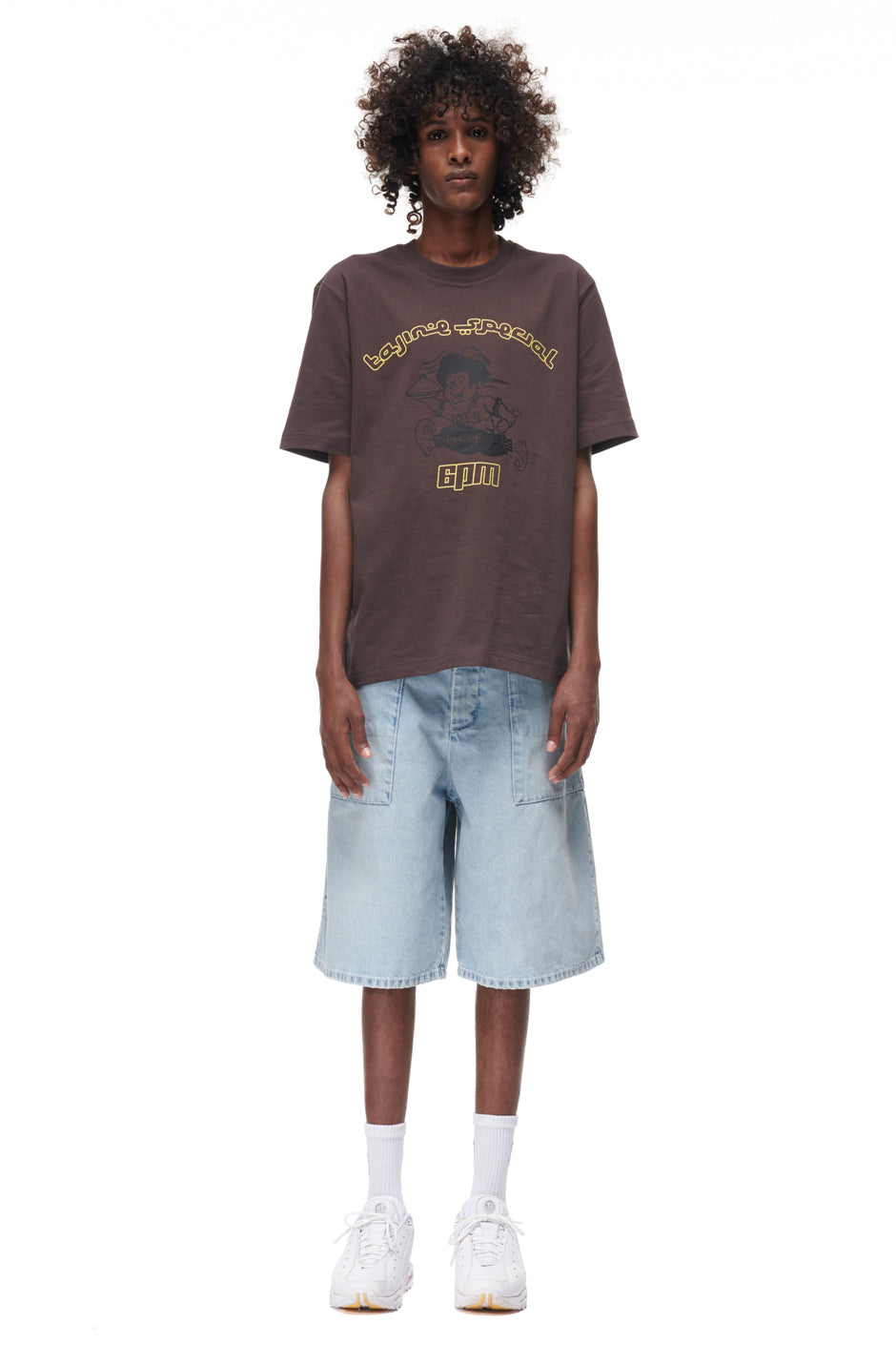 A person with curly hair wears the 6PM TAJIN T-SHIRT BROWN, light blue knee-length denim shorts, white socks, and white sneakers, standing against a plain white background.
