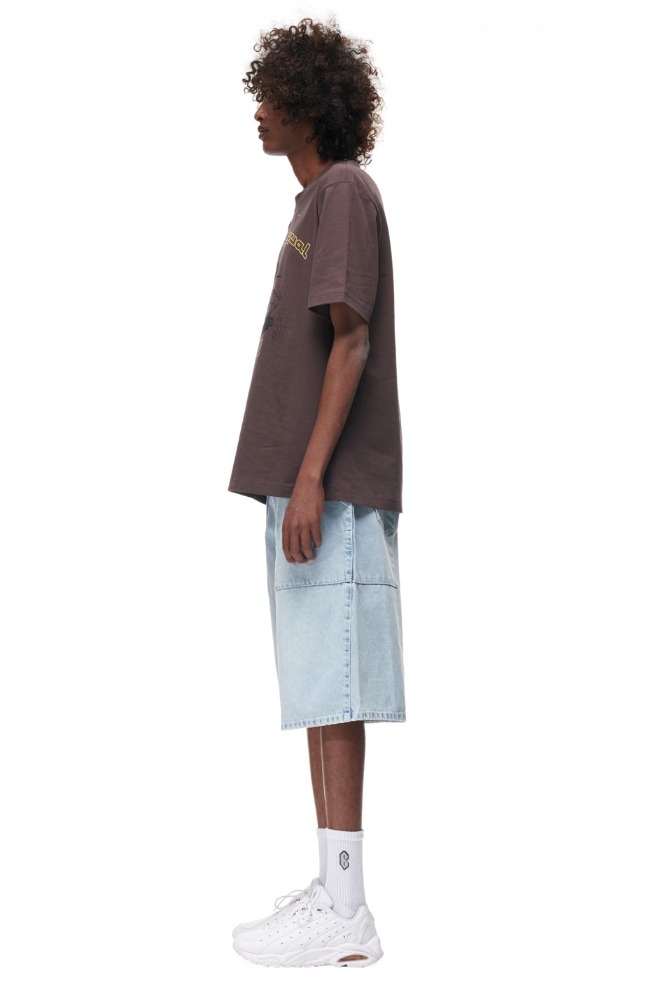A person with curly hair stands in profile against a white background, wearing the 6PM TAJIN T-SHIRT BROWN, light blue knee-length denim shorts, white crew socks, and white sneakers.