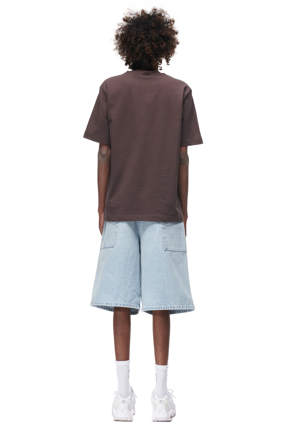 A person with curly hair faces away, wearing the 6PM TAJIN T-SHIRT BROWN, light blue knee-length denim shorts with large pockets, white socks, and white sneakers, all set against a plain white background.