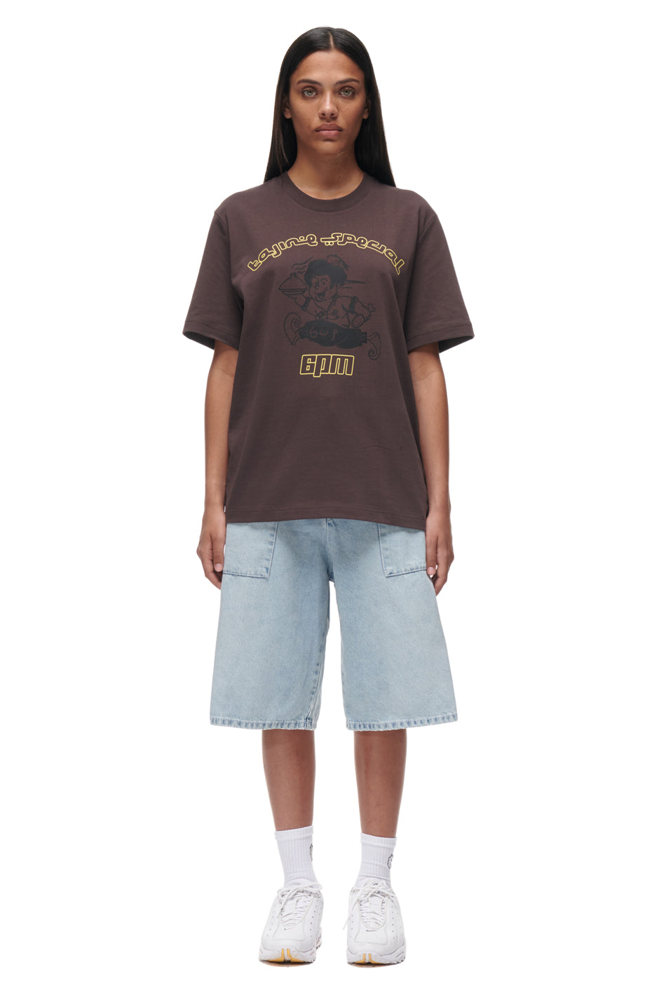 A woman stands facing forward against a plain white background, wearing the 6PM TAJIN T-SHIRT BROWN styled with light blue knee-length denim shorts, white crew socks, and white sneakers.