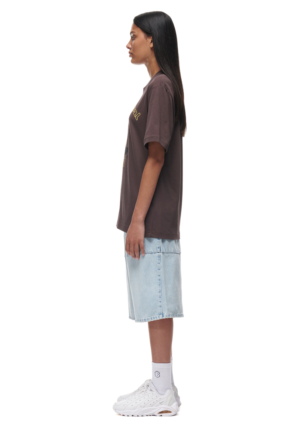 A person with long dark hair stands in profile, wearing the 6PM TAJIN T-SHIRT BROWN, light blue knee-length denim shorts, white crew socks, and white sneakers against a plain white background.