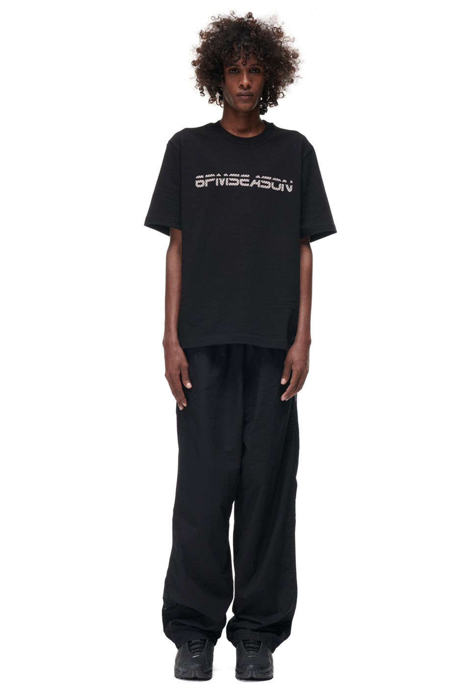 A person with curly hair stands facing forward against a white background, wearing a black 6PM TILES T-SHIRT, loose black pants, and black sneakers.