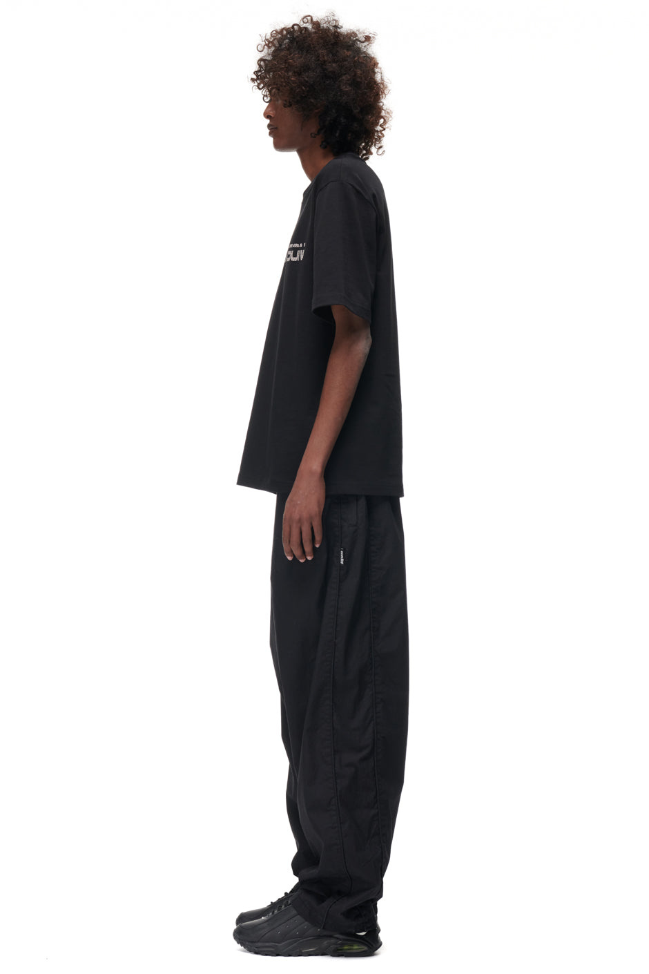 A person with curly hair stands in profile against a white background, wearing the 6PM TILES T-SHIRT BLACK, paired with loose black pants and black sneakers.