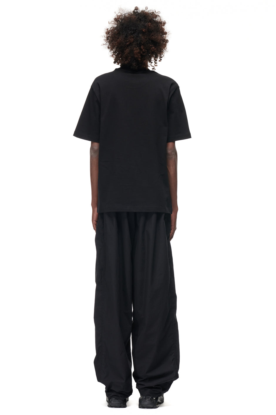 A person with curly hair faces away, wearing the 6PM TILES T-SHIRT BLACK, loose black pants, and black shoes against a plain white background.