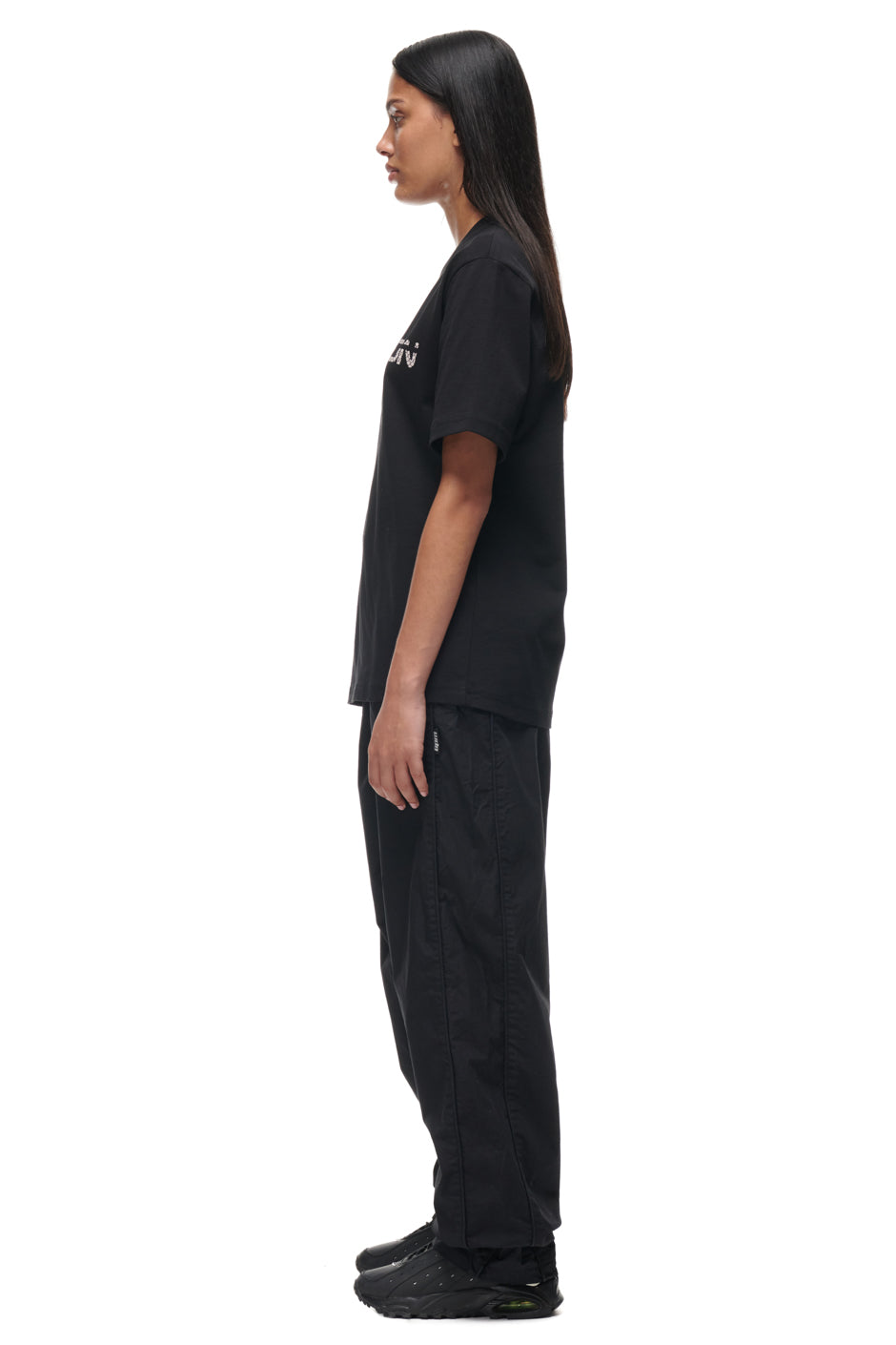 A woman with long, straight dark hair stands in profile against a white background, wearing the TILES T-SHIRT BLACK by 6PM, loose black pants, and black sneakers, with her arms resting naturally by her sides.