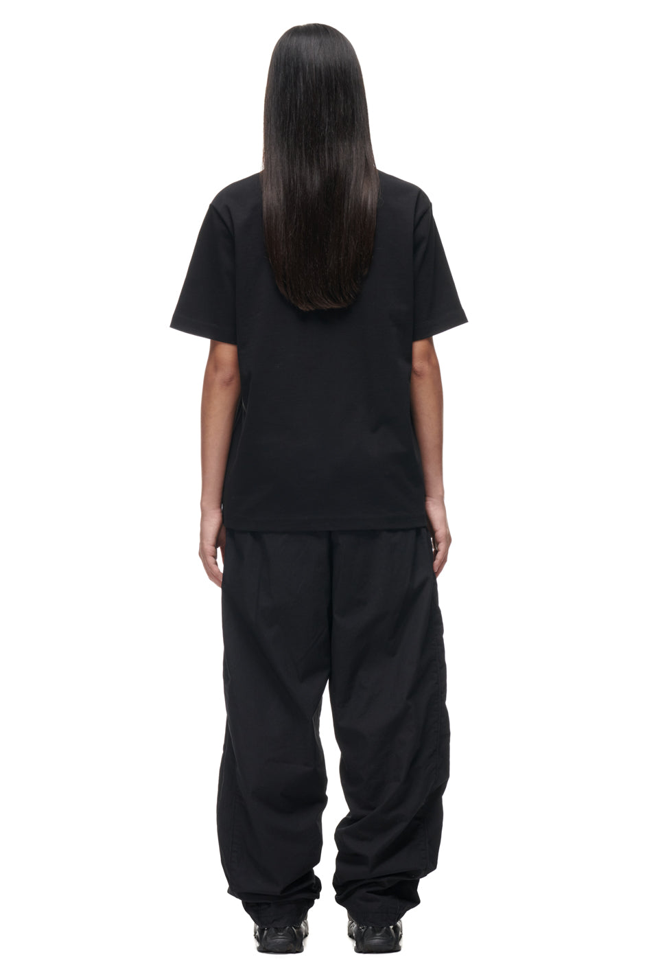 A person with long, straight dark hair stands facing away against a plain white background, wearing the 6PM TILES T-SHIRT BLACK, paired with baggy black pants and black shoes.