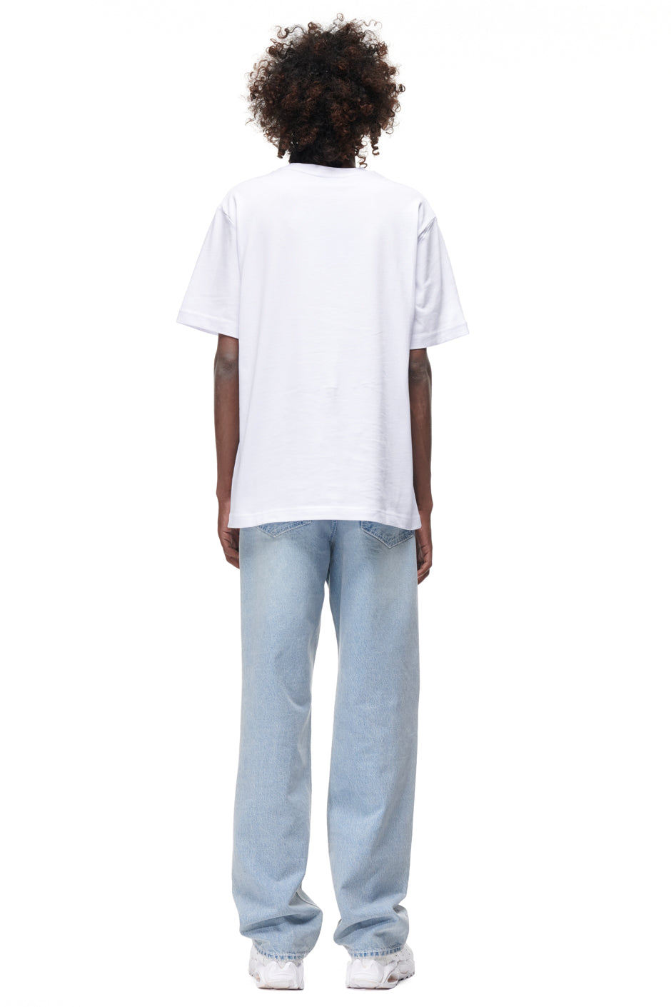 A person with curly hair faces away from the camera, wearing the 6PM TAJIN T-SHIRT WHITE, light blue jeans, and white sneakers against a white background.