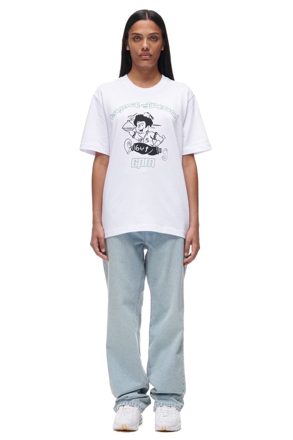 A person with long dark hair stands facing forward against a white background, wearing the 6PM TAJIN T-SHIRT WHITE featuring cartoon art, paired with light blue jeans and white sneakers.