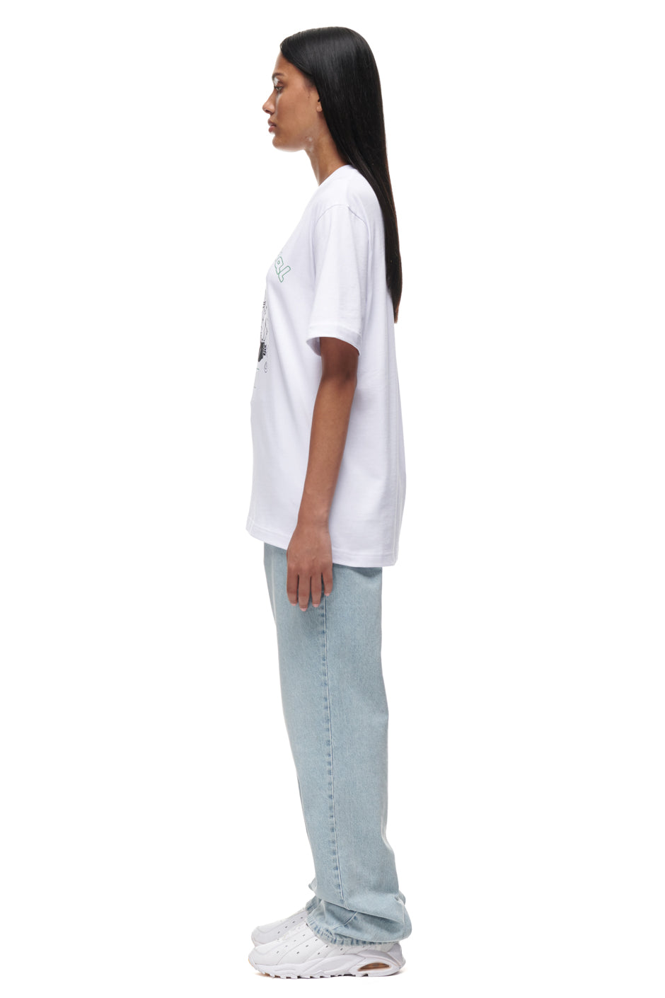 A woman with straight black hair stands in profile against a white background, wearing the 6PM TAJIN T-SHIRT WHITE, light blue jeans, and white sneakers.