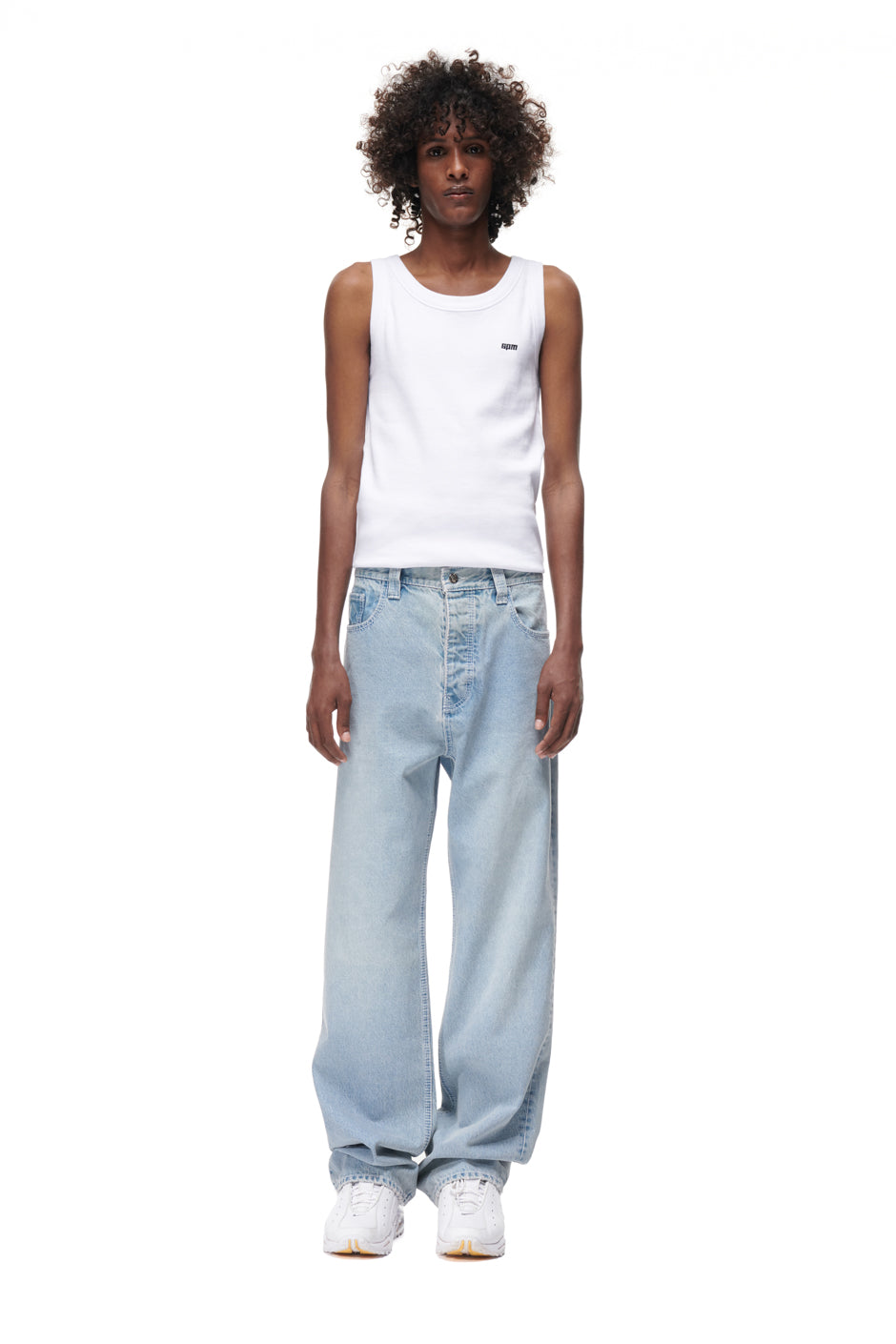 A person with curly hair wears 6PM's DENIM JEANS, a white tank top, and white sneakers, standing facing forward against a plain white background.