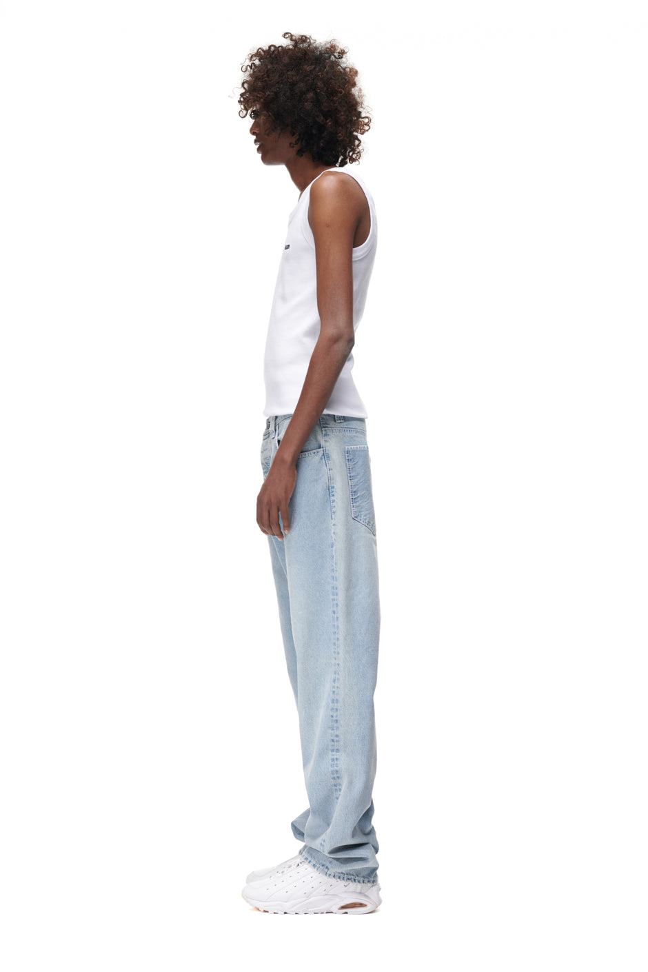 A person with curly hair stands in profile against a white background, wearing a white tank top, 6PM DENIM JEANS, and white sneakers.
