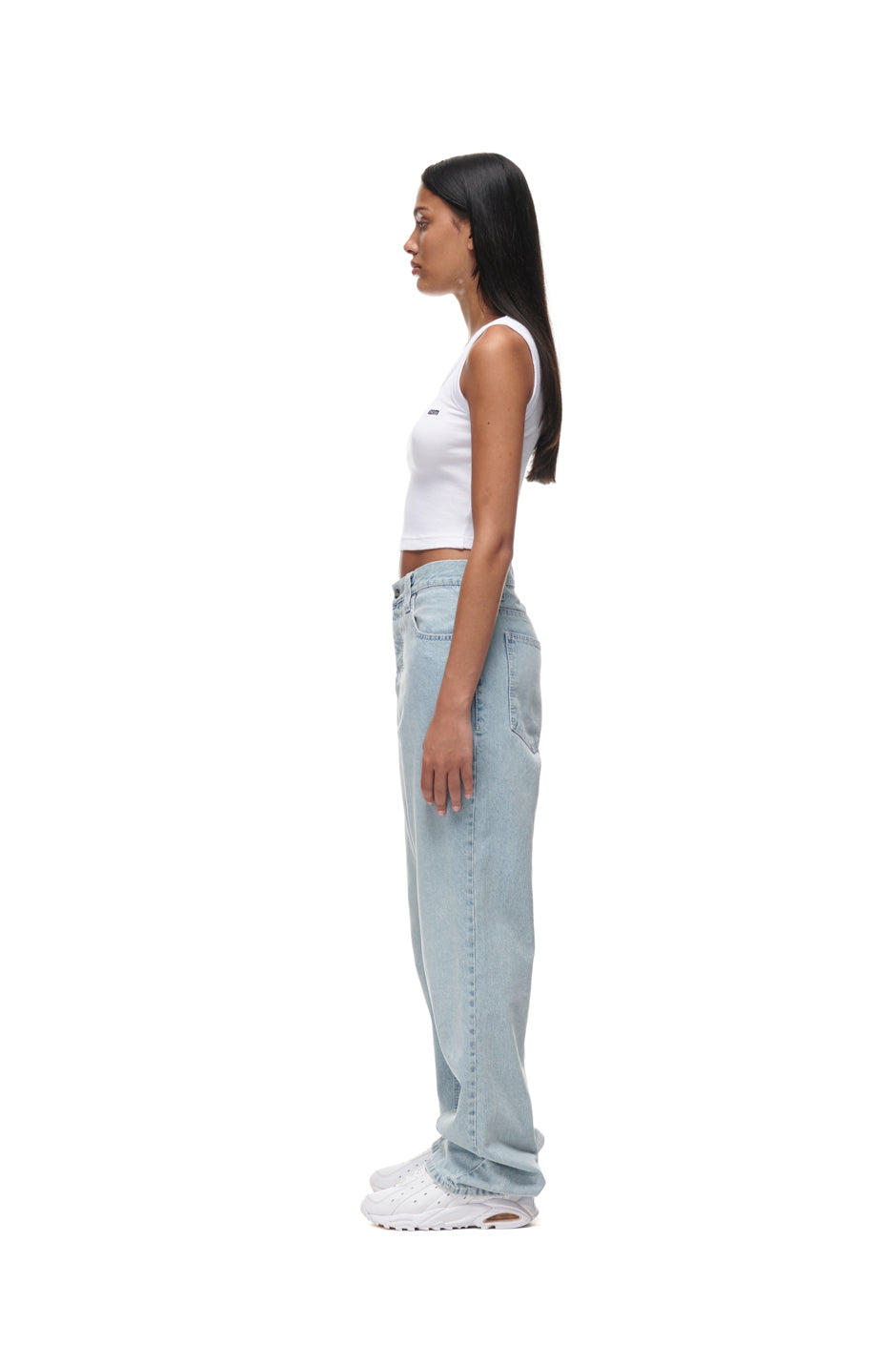 A woman with long dark hair stands in profile against a white background, wearing a white sleeveless crop top, 6PM DENIM JEANS, and white sneakers.