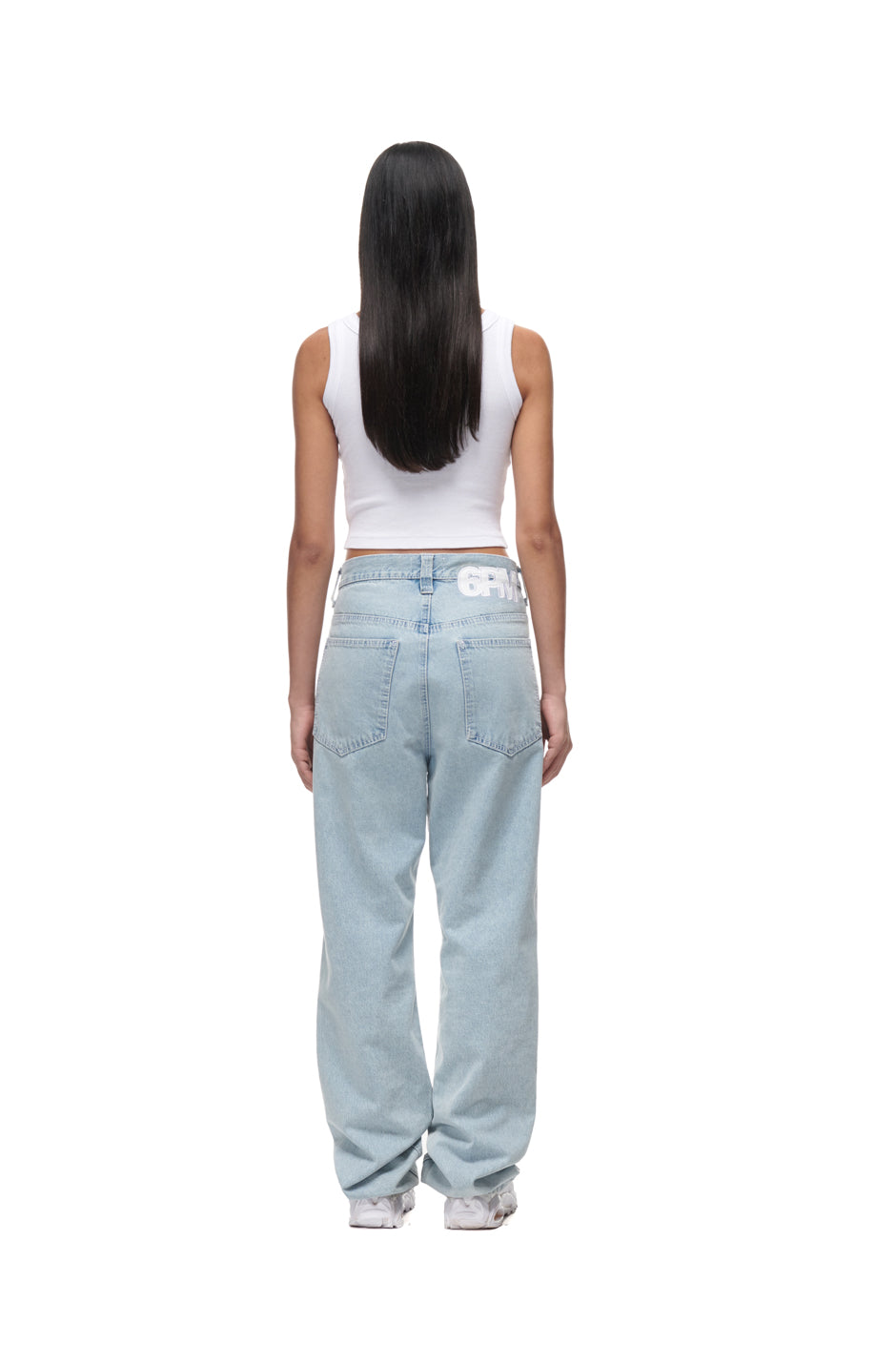 A person with long dark hair, facing away, is wearing 6PM DENIM JEANS with a white sleeveless crop top and white sneakers, standing against a plain white background.