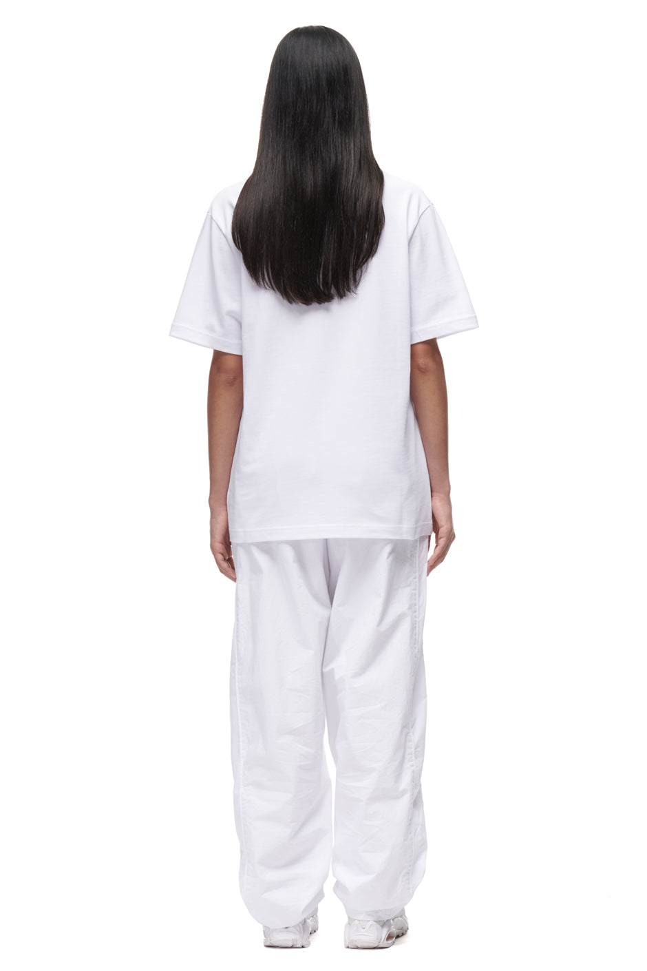 A person with long, straight dark hair faces away, wearing the 6PM TILES T-SHIRT WHITE, paired with white pants and shoes, set against a plain white background.
