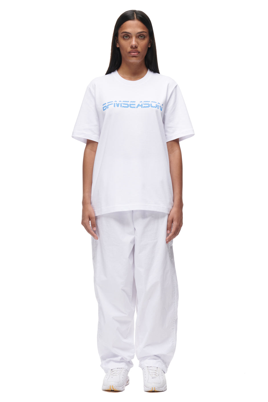 A woman faces forward against a plain white background, wearing the 6PM TILES T-SHIRT WHITE with blue "8PM SEASON" text, wide white pants, and white sneakers.