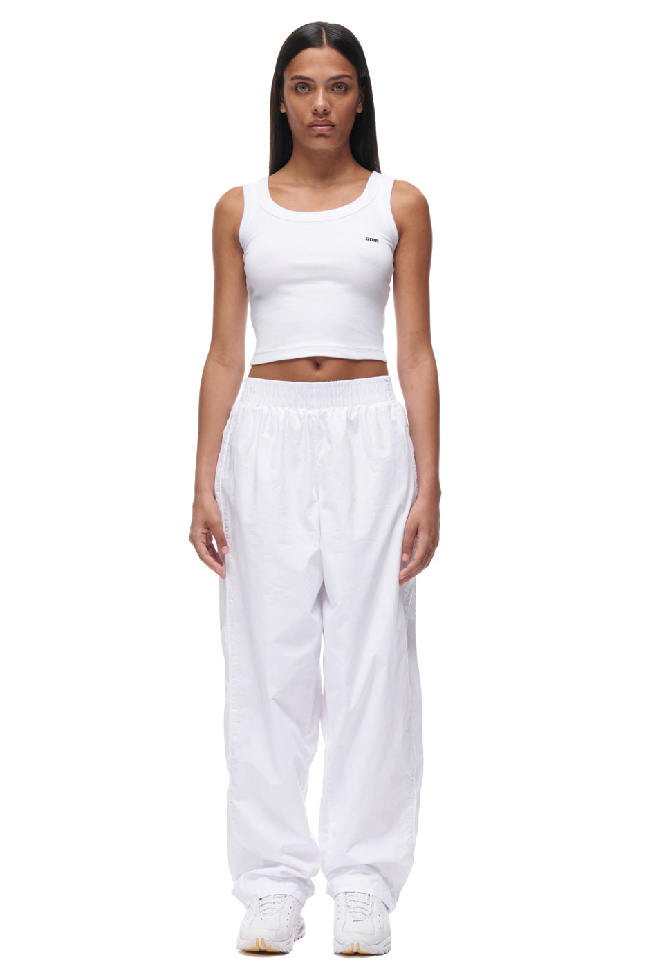 A woman with straight dark hair and a neutral expression stands facing forward against a plain white background, wearing 6PM TRACKPANTS WHITE, a fitted white crop tank top, and white sneakers. Her arms are relaxed by her sides.