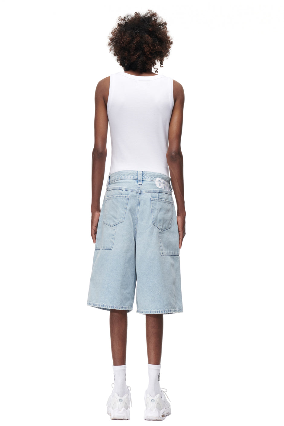 A person stands with their back to the camera, wearing 6PM DENIM SHORTS, a white tank top, white socks, and white sneakers, all set against a plain white background.