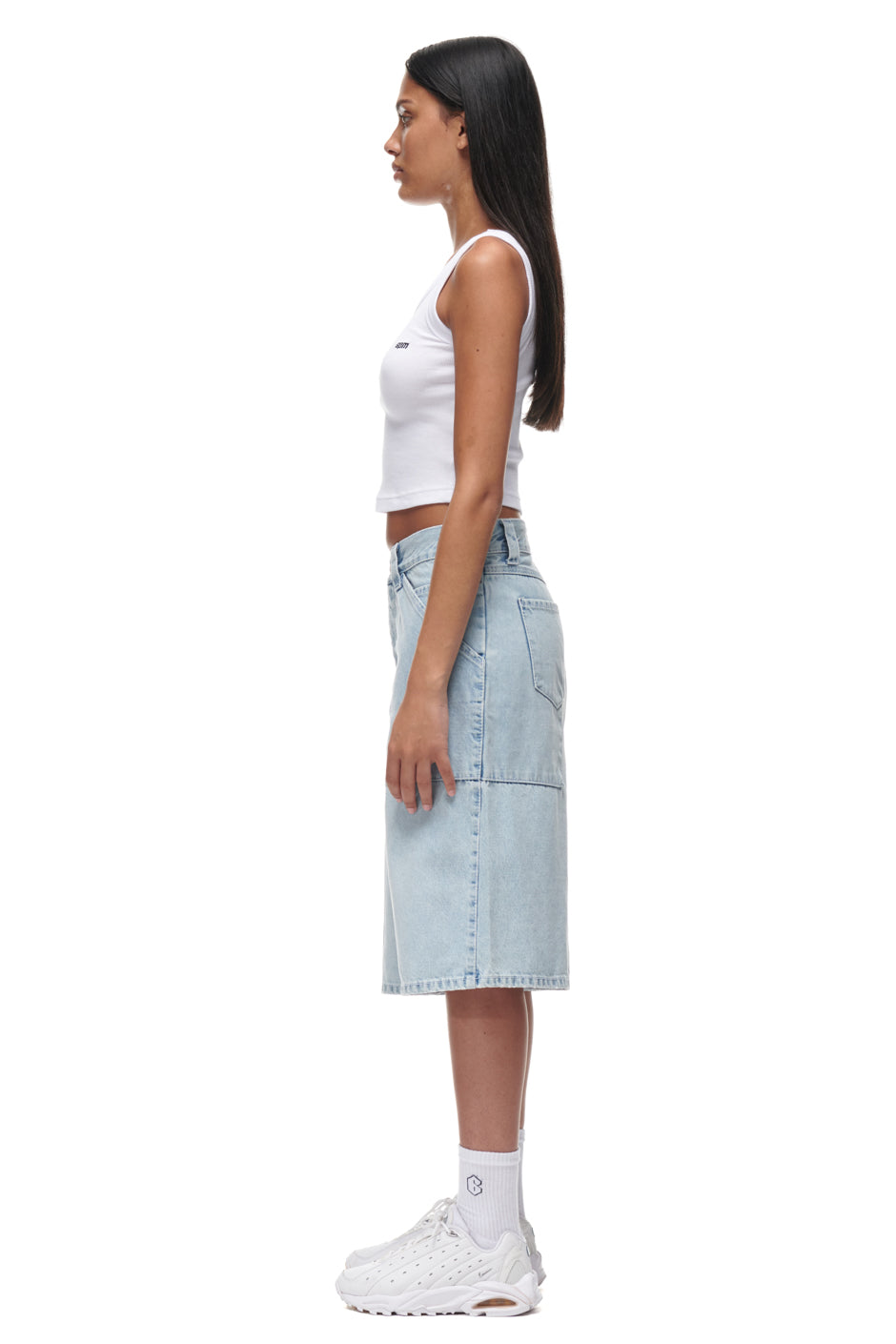 A woman in profile, wearing 6PM DENIM SHORTS, a white sleeveless crop top, white socks, and white sneakers against a white background. She has long straight dark hair.