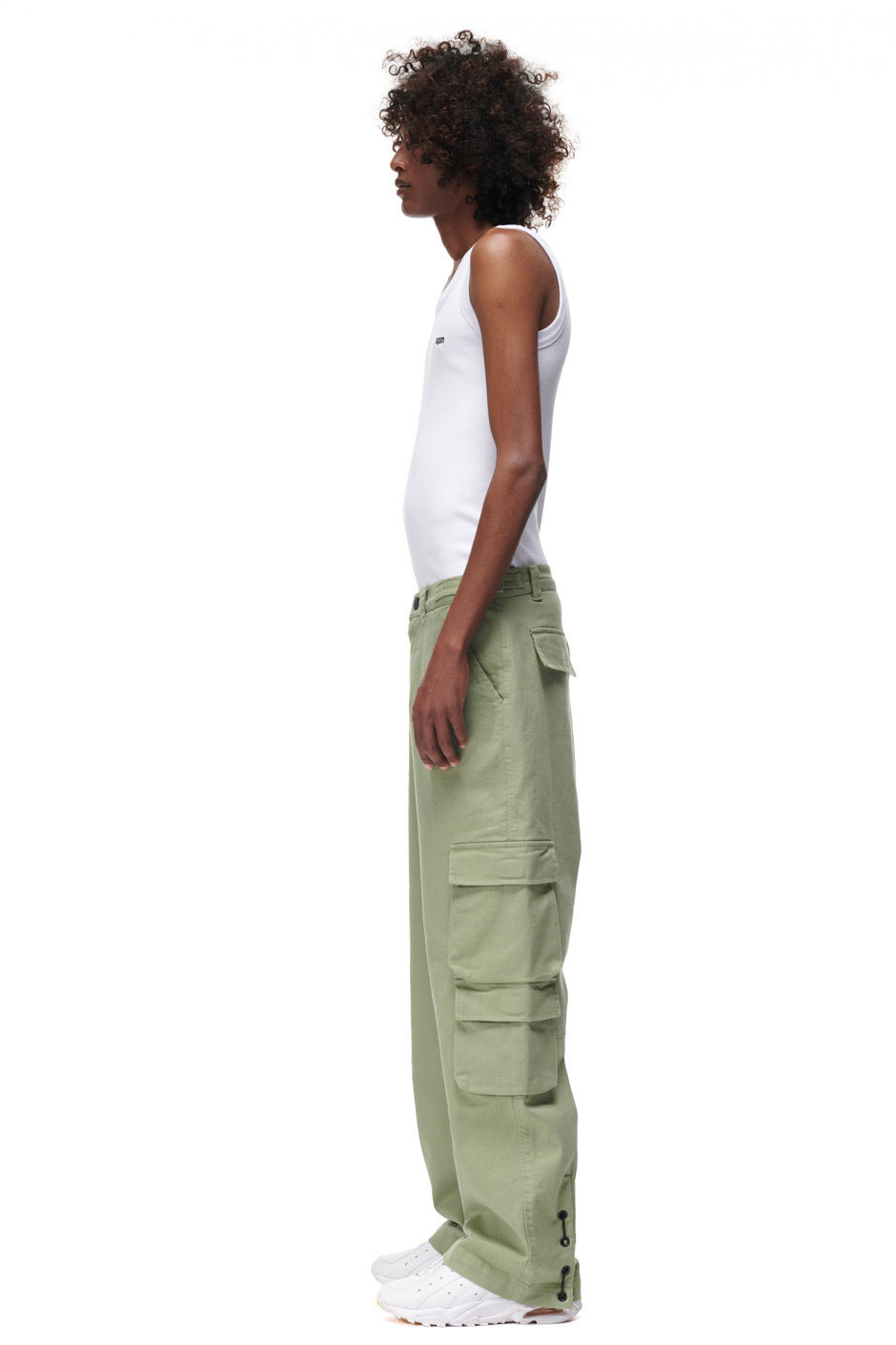 A person with curly hair in a white tank top, 6PM's CARGO PANTS GREEN, and white sneakers stands in profile against a white background.