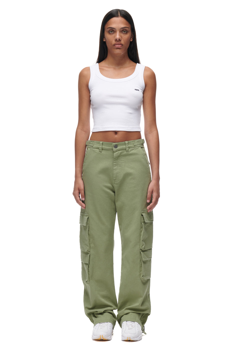 A woman with long straight hair wears 6PM's CARGO PANTS GREEN, paired with a white crop tank top and white sneakers. She stands facing forward with a neutral expression against a plain white background.