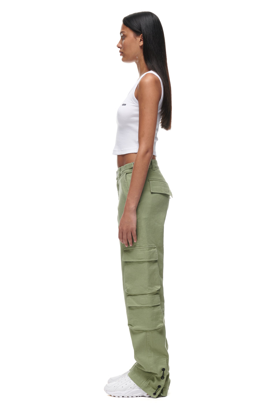 A woman stands in profile against a white background, wearing a white sleeveless crop top, 6PM CARGO PANTS GREEN with pockets, and white sneakers. Her long dark hair is straight and loose, arms resting by her sides.