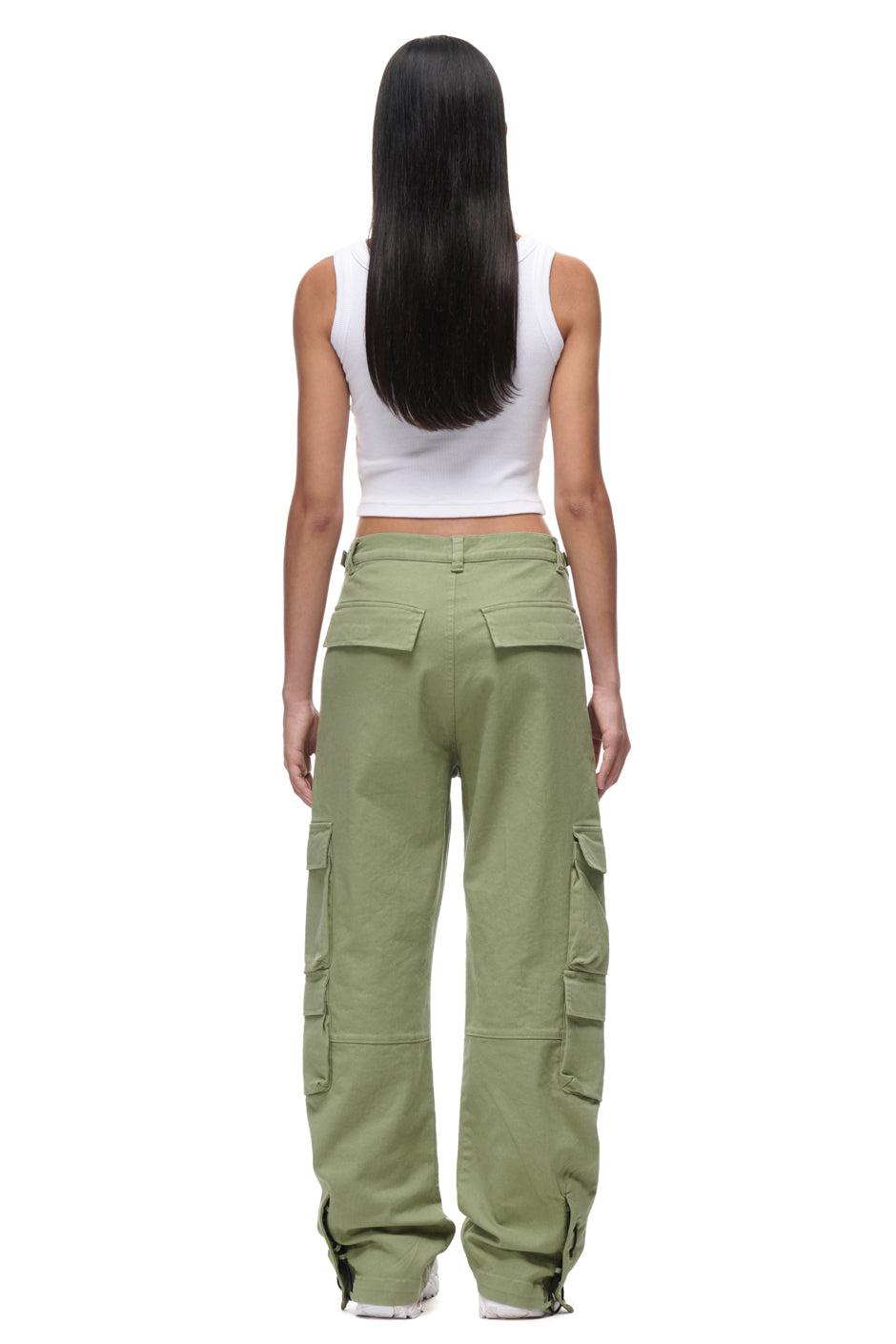 A person with long straight hair stands facing away, wearing a white sleeveless crop top and 6PM CARGO PANTS GREEN—loose-fitting, light green pants with pockets. The background is white.