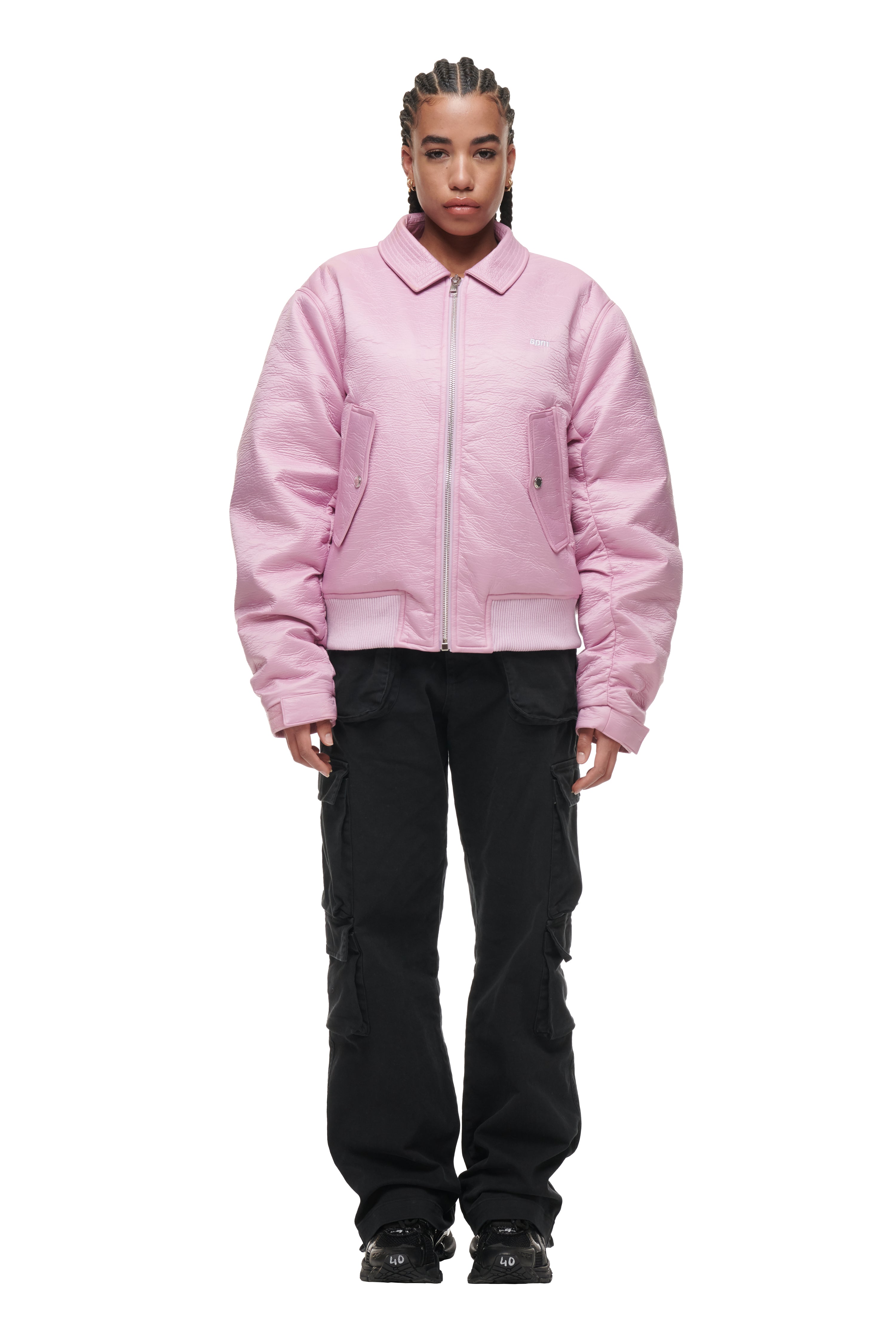 A person with cornrow braids models the 6PM BOMBER JACKET PINK, paired with black cargo pants and black shoes, standing facing forward against a white background.