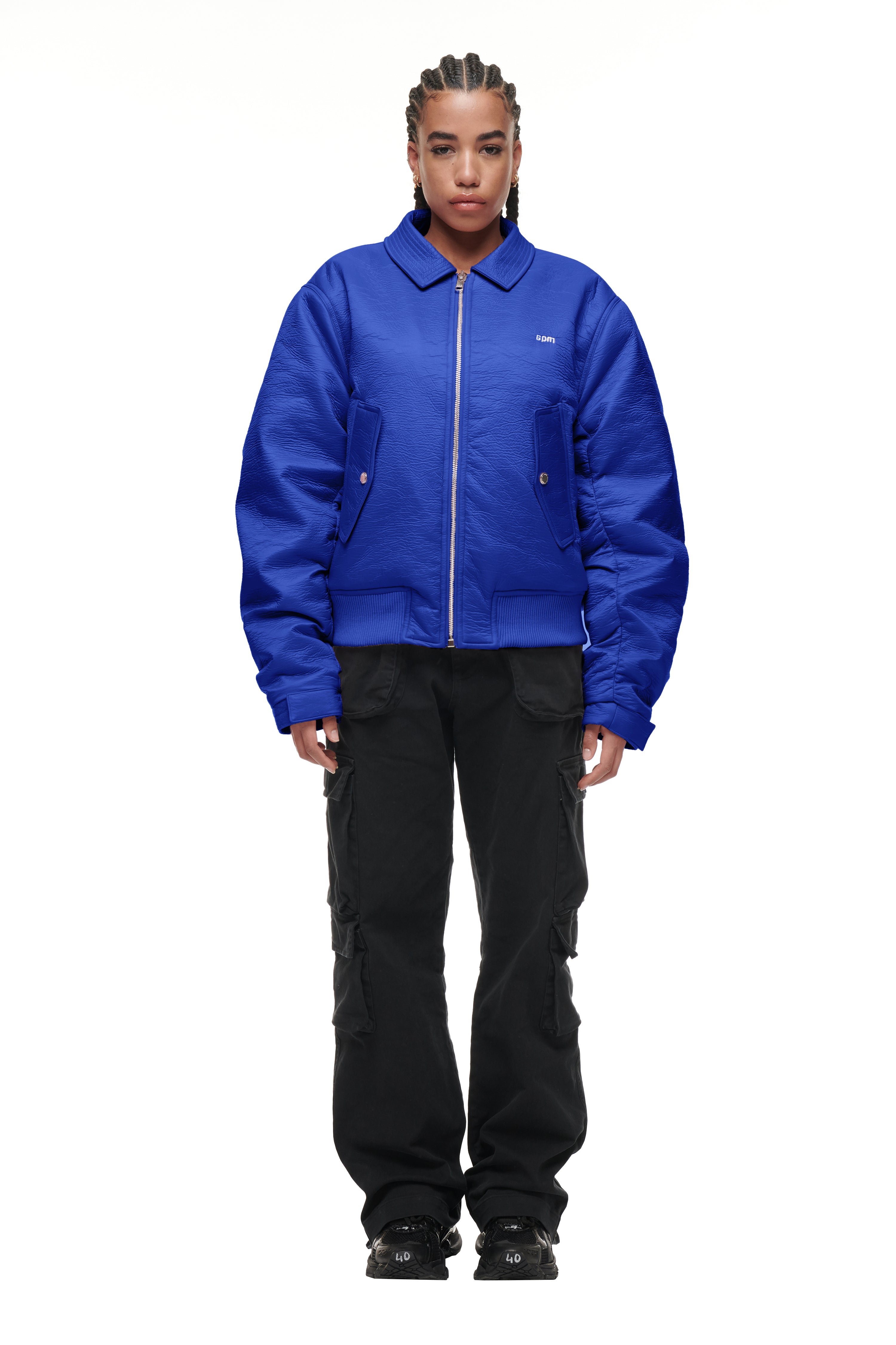 A person with braids faces forward, wearing the 6PM BOMBER JACKET BLUE with black cargo pants and black shoes, standing against a plain white background.