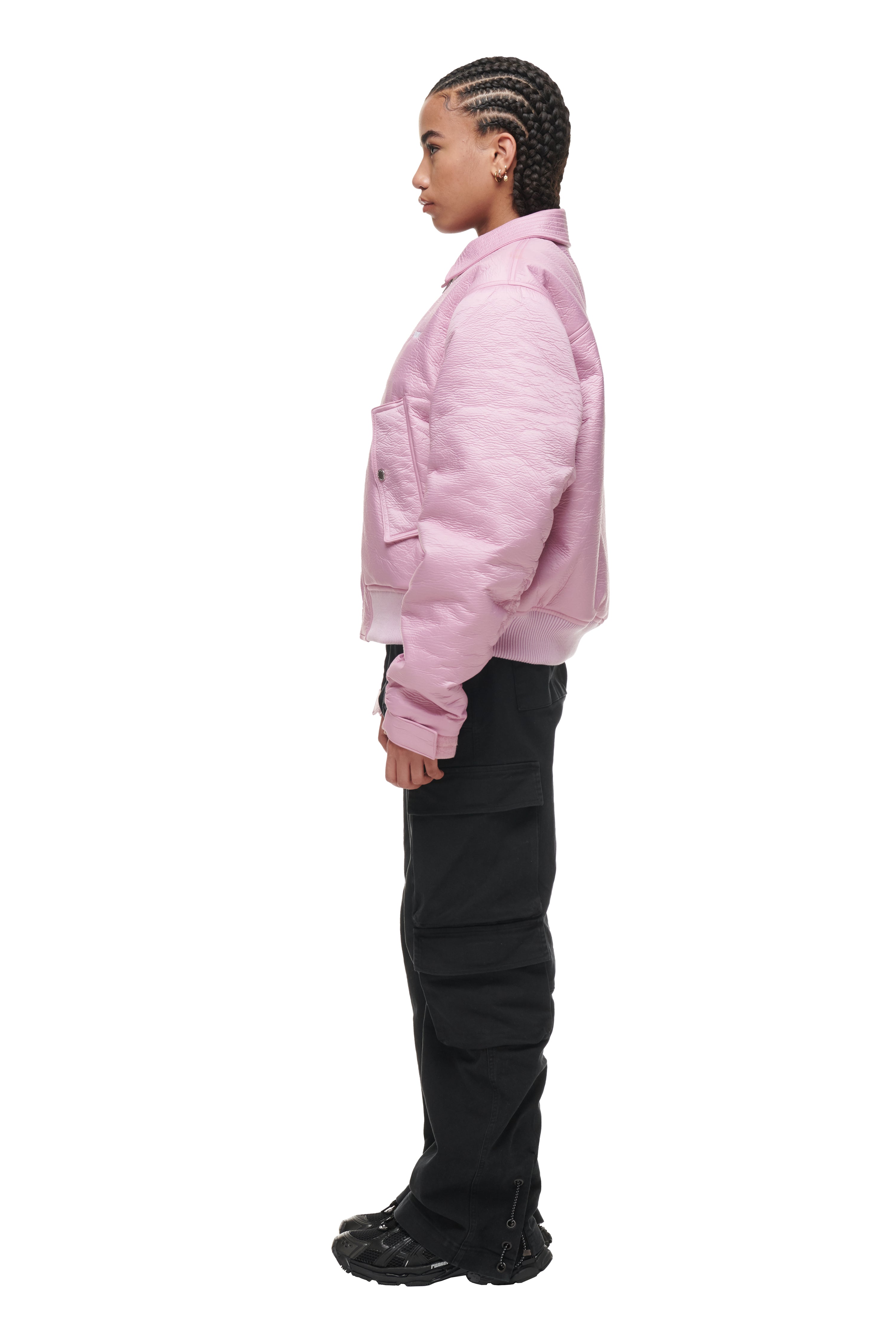 A person with braided hair stands in profile against a white background, wearing the 6PM BOMBER JACKET PINK, paired with black cargo pants and black sneakers.