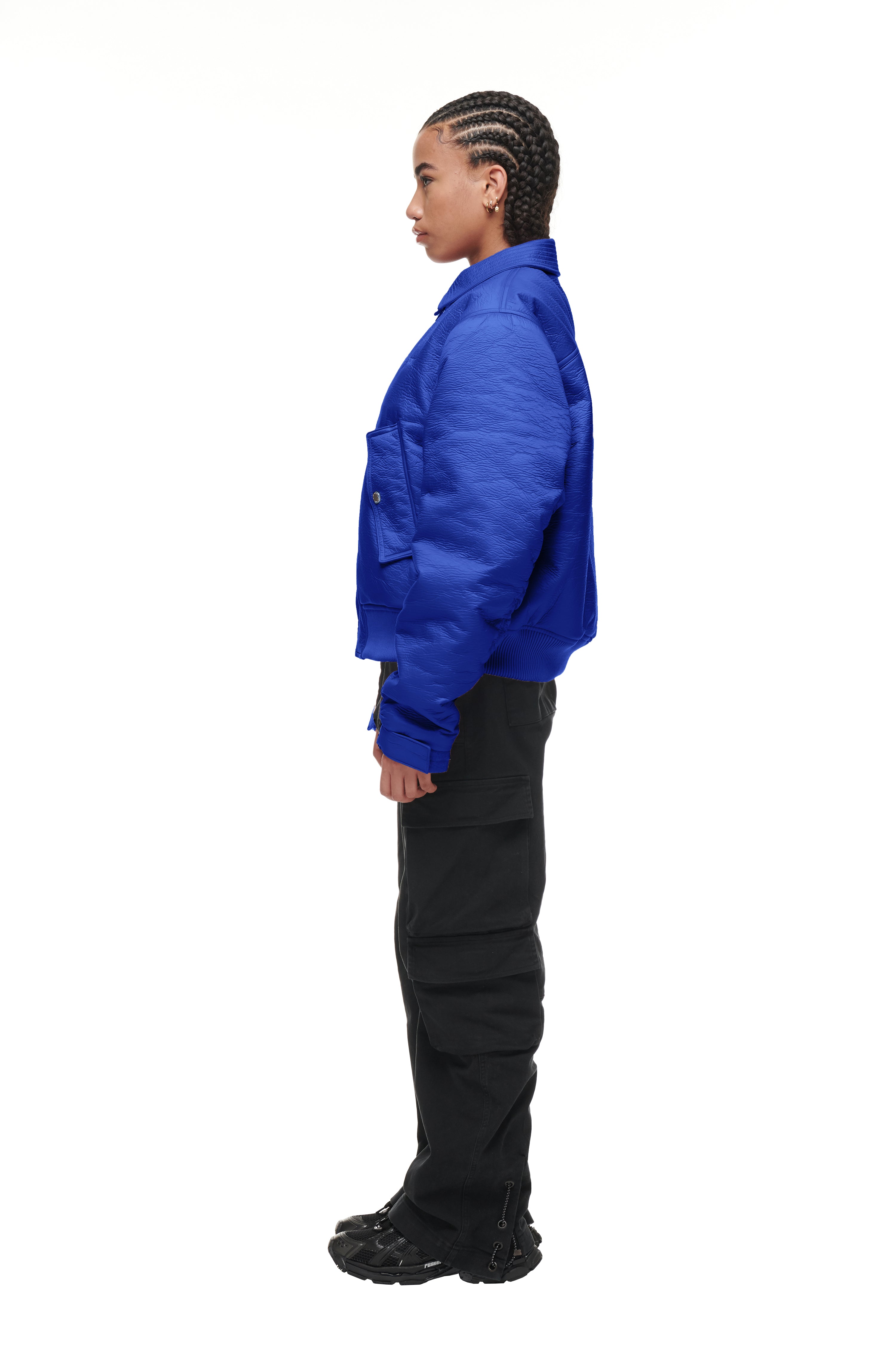 A person with braided hair stands in profile against a white background, wearing the 6PM BOMBER JACKET BLUE, black cargo pants, and black shoes.