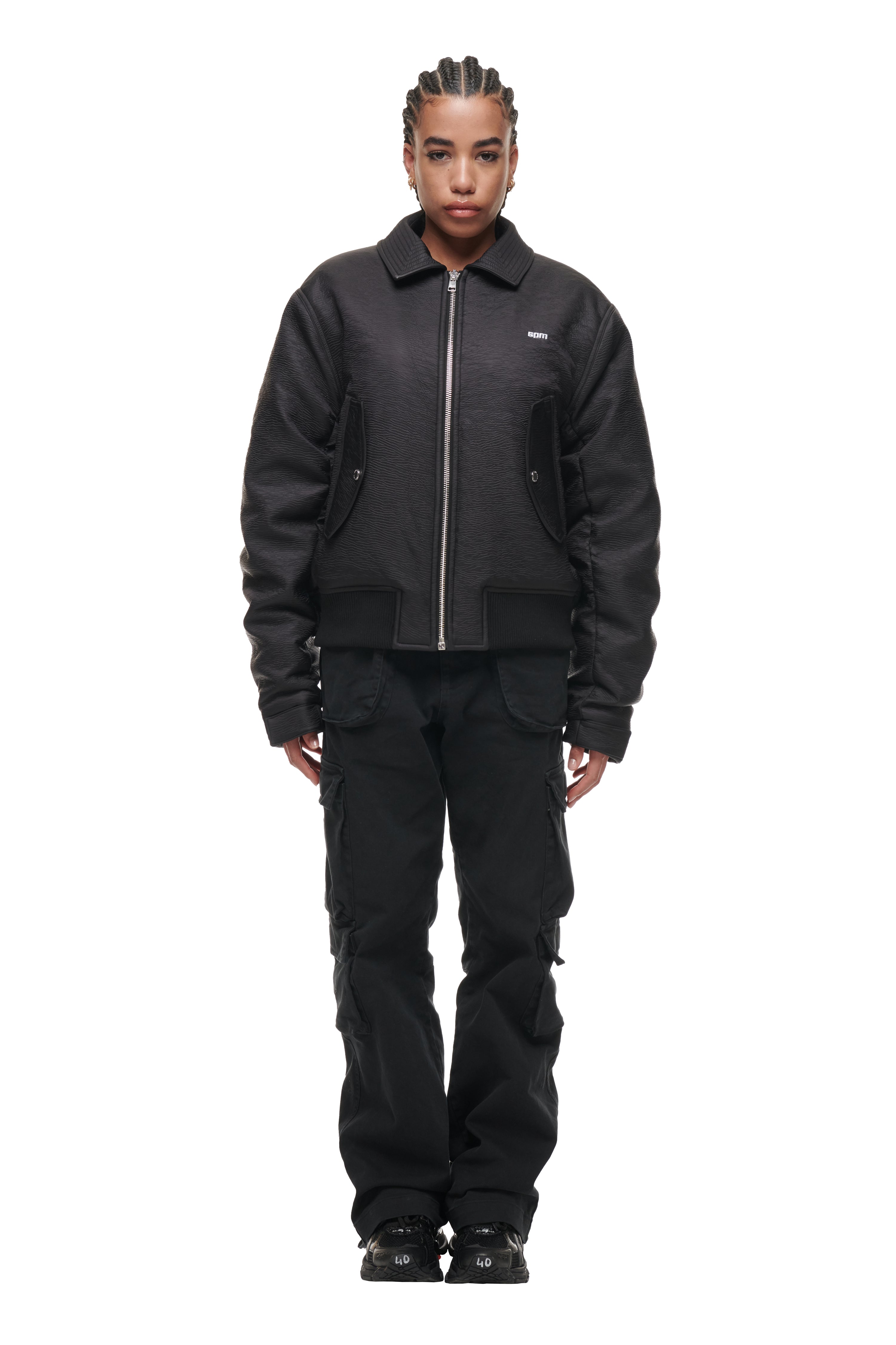 A person faces forward against a white background, wearing the 6PM BOMBER JACKET BLACK, black cargo pants, and black shoes. Their hair is styled in tight braids.