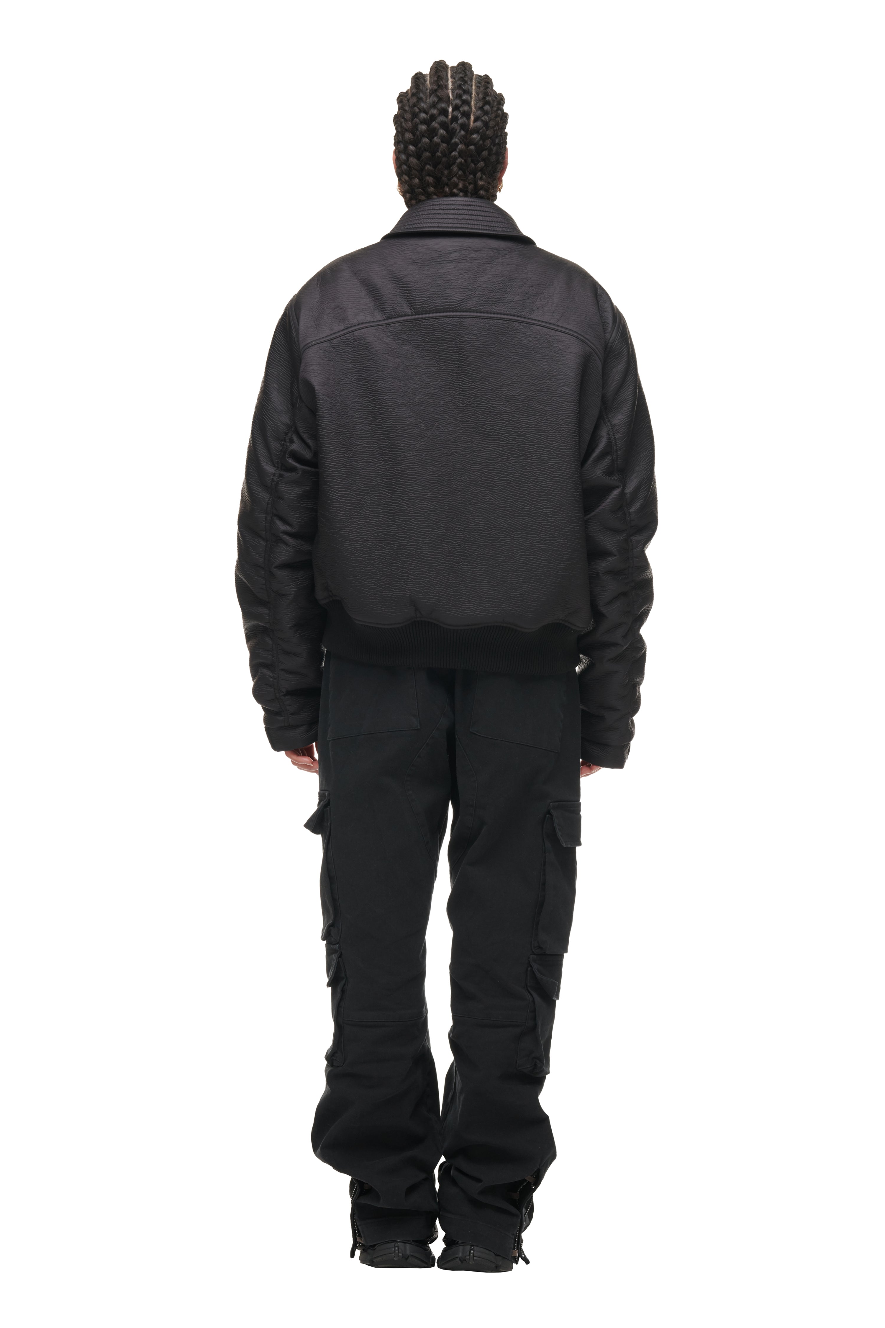A person with neat braids faces away from the camera, wearing a 6PM BOMBER JACKET BLACK, black cargo pants, and black shoes against a plain white background.